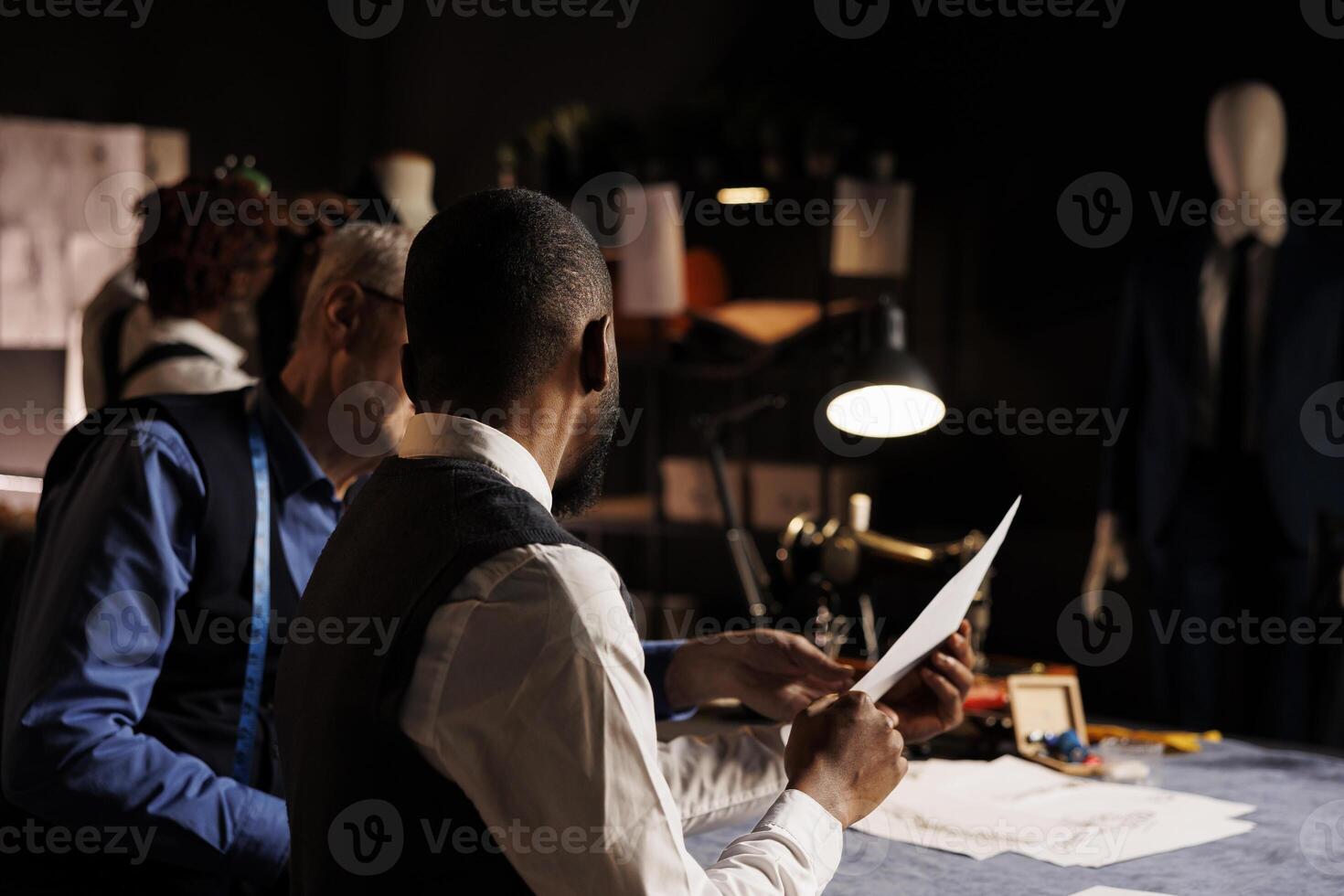 African american client discussing over clothing sketches with skilled craftsman, asking for customization alterations to be done. Suitmaker helping gentleman with bespoke attire in atelier studio photo