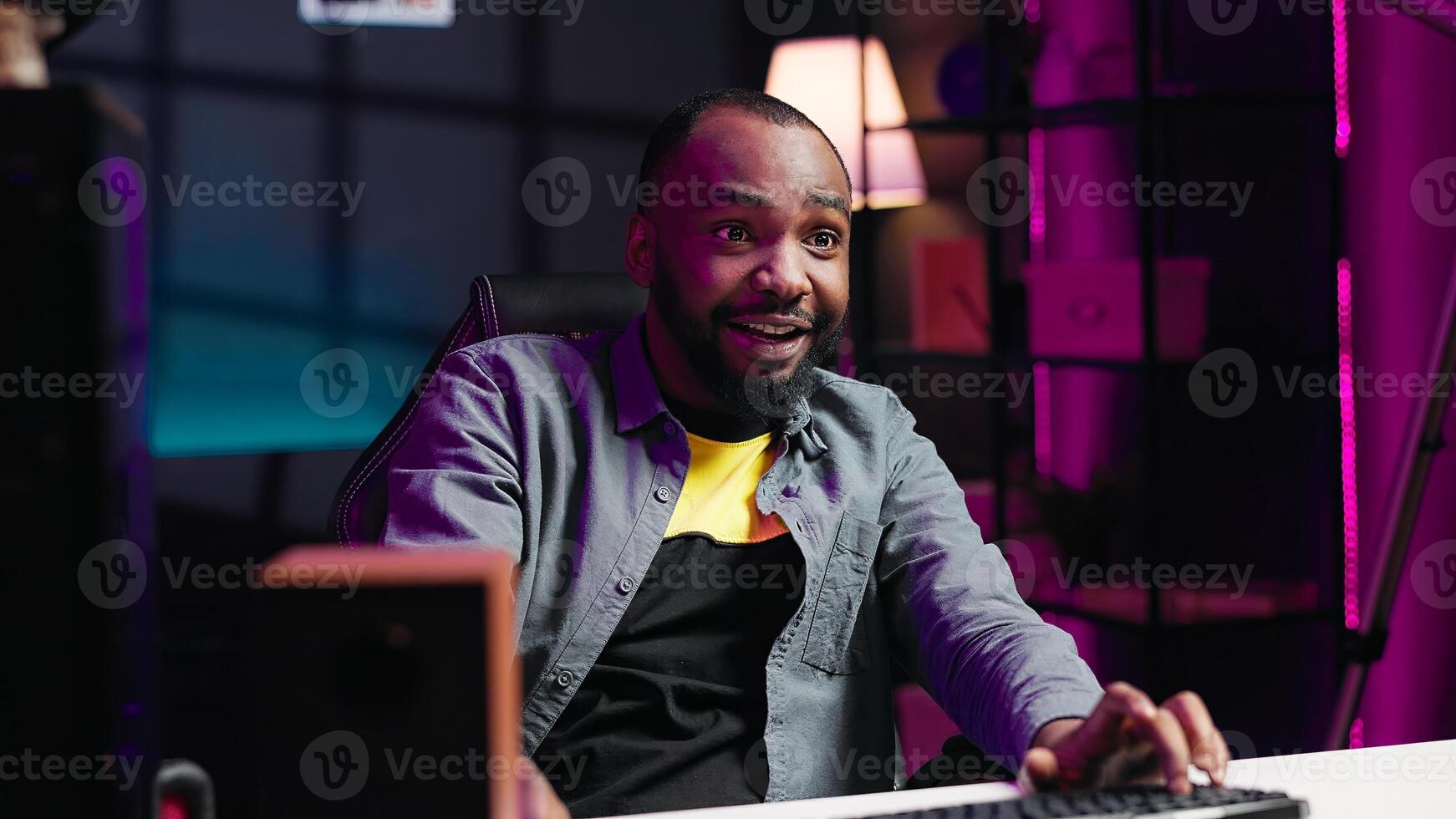 Joyous gamer competing in intense internet multiplayer videogame from vibrant living room with neon lights. Smiling african american man using PC, having immersive experience while gaming, camera A photo
