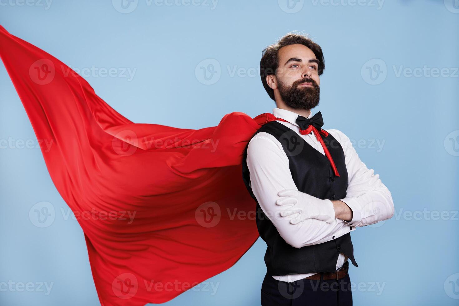 Strong man hero with red mantle standing against blue background, wearing superhuman cartoon costume on camera. Male person showing force and strength to defend people, superhero. photo