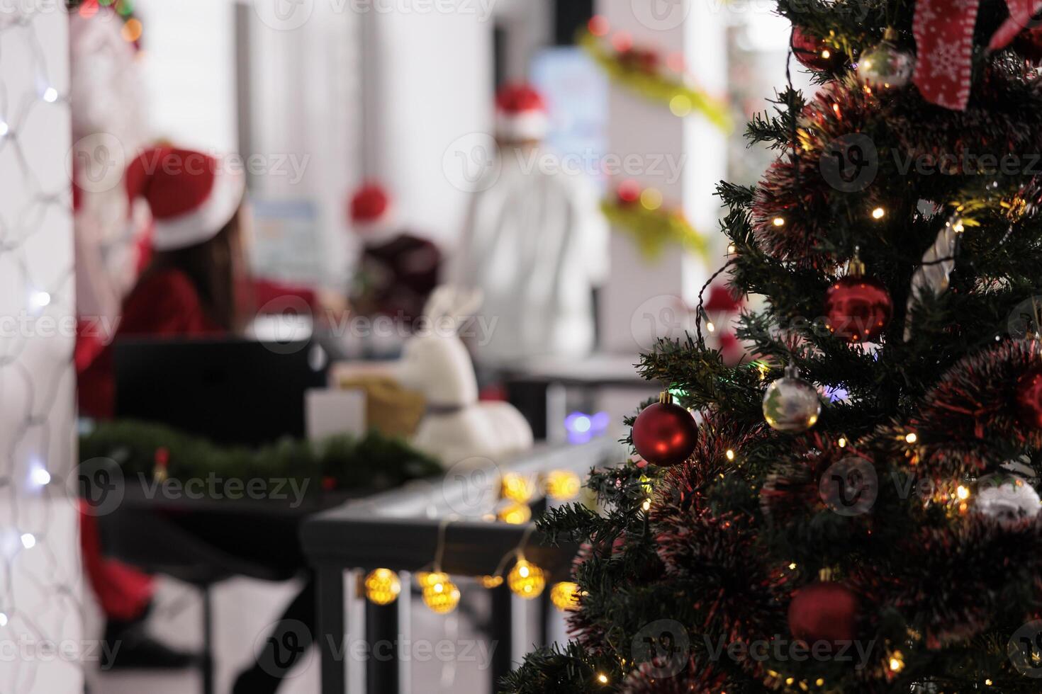 Stylishly decorated Christmas tree close up with employees receiving special gifts from Santa Claus on winter holidays in blurred background. Xmas costumed manager giving red box presents to workers photo