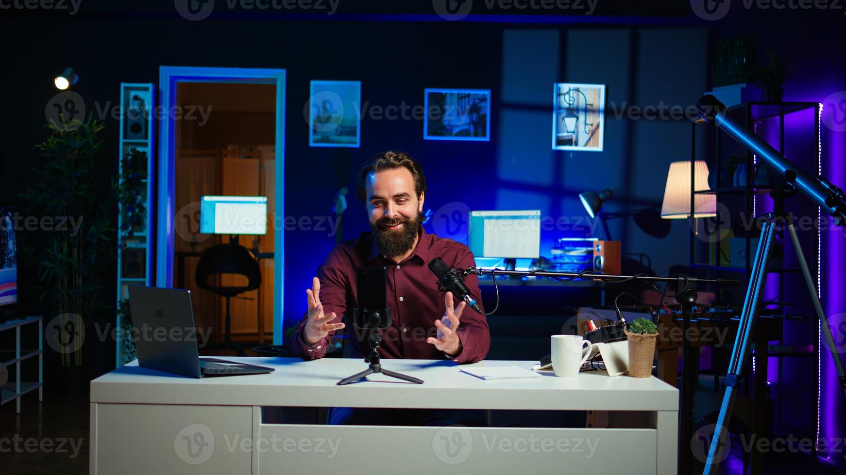 Content creator adjusting professional microphone in studio before starting vlog, recording with selfie camera. Man in neon lit home studio preparing equipment, filming with smartphone, camera B photo