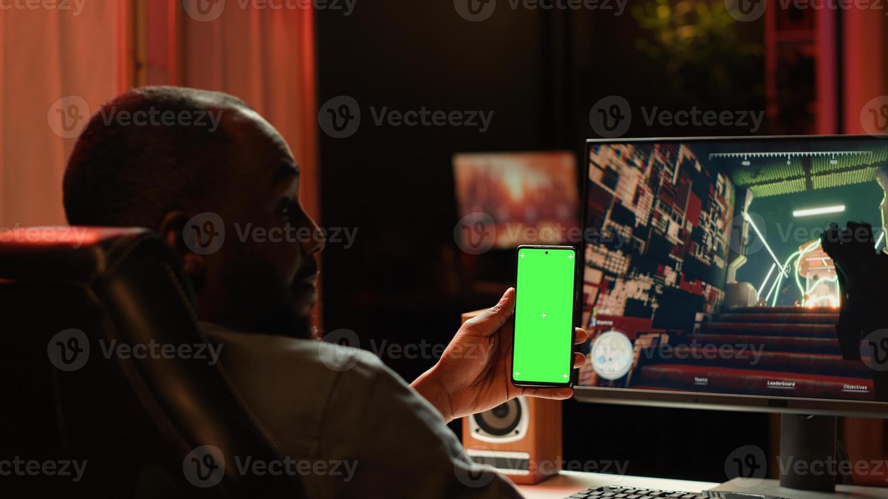 Isolated screen smartphone held by player competing in online multiplayer videogame at home. Mockup mobile phone utilized by person playing on gaming PC in neon illuminated apartment photo