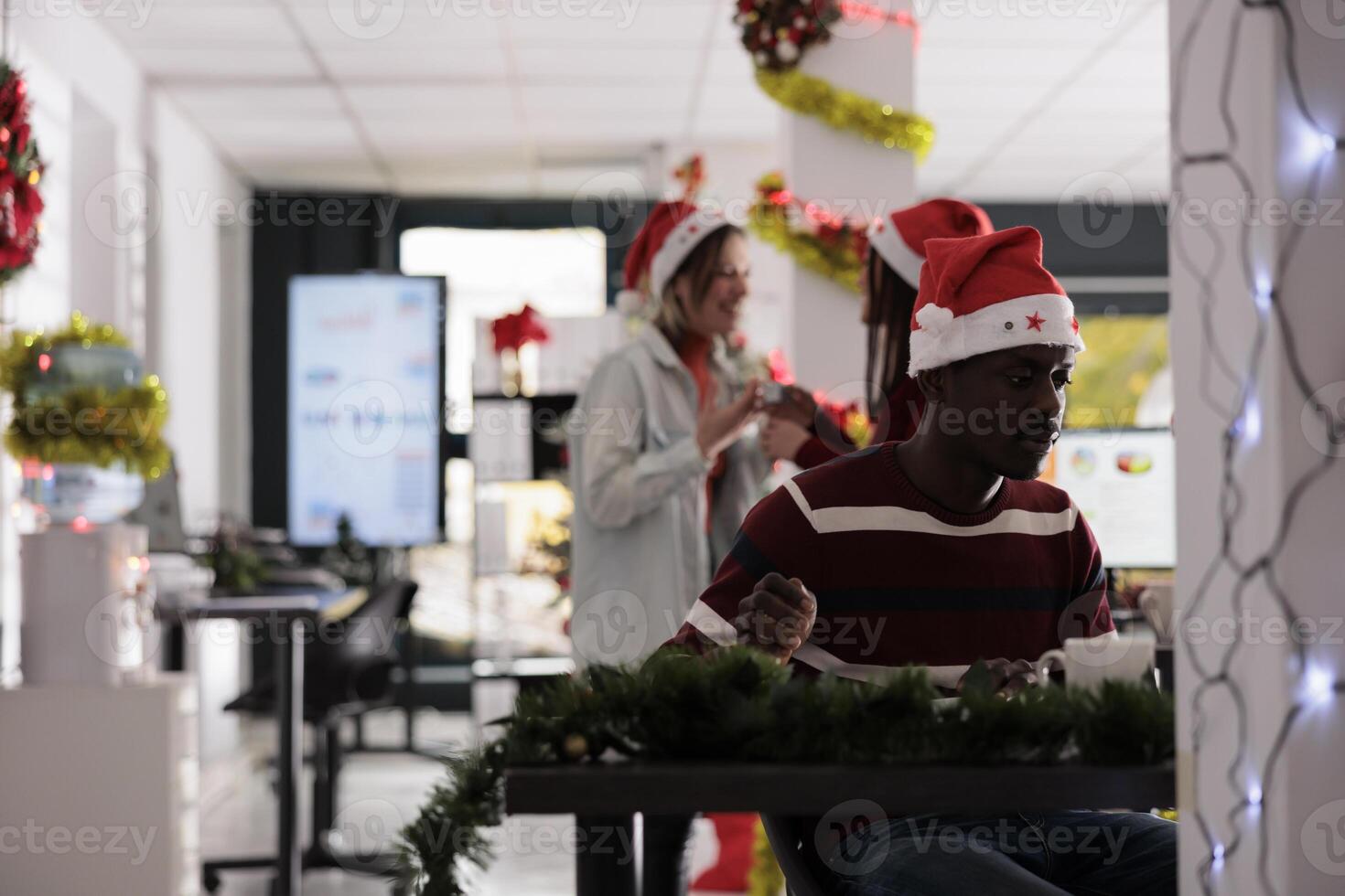 Employee wearing Christmas themed clothing focused on finishing tedious paperwork while coworkers have fun in background. Exhausted african american worker solving tasks in festive decorated office photo