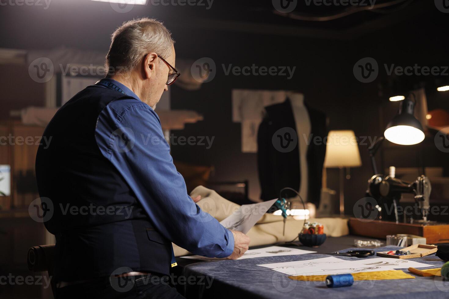 Elderly couturier looking over sketch drawings, preparing to create custom made sartorial garments for comissioning client. Suitmaker conceptualizing outfits following handdrawn drafts photo