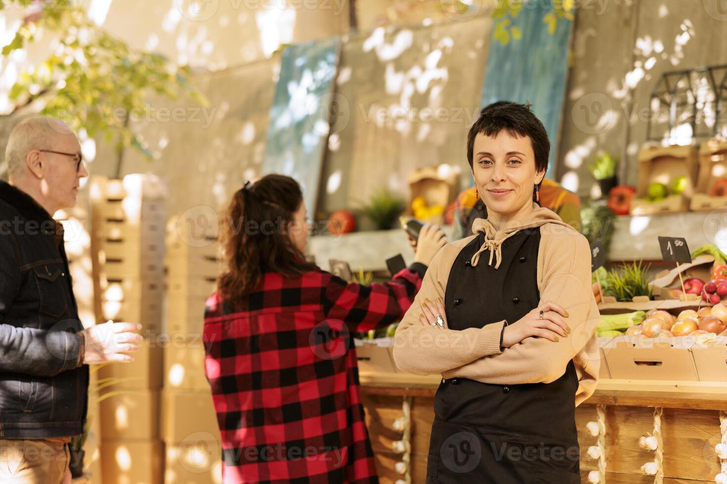 Caucasian woman in an apron runs a farmers market stall, selling fresh organic produce directly to customers. Female greengrocer standing with her arms crossed and looking at the camera. photo