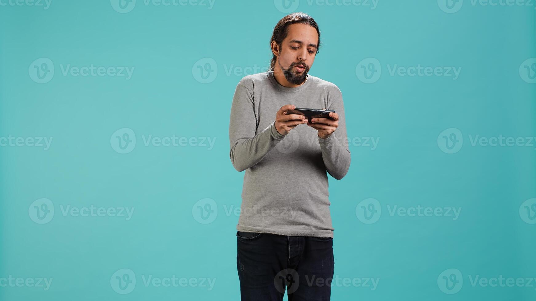 Focused man plays intense videogame on mobile phone, spamming attack buttons on touchscreen. Gamer participates in gaming competition, isolated over studio background, camera A photo