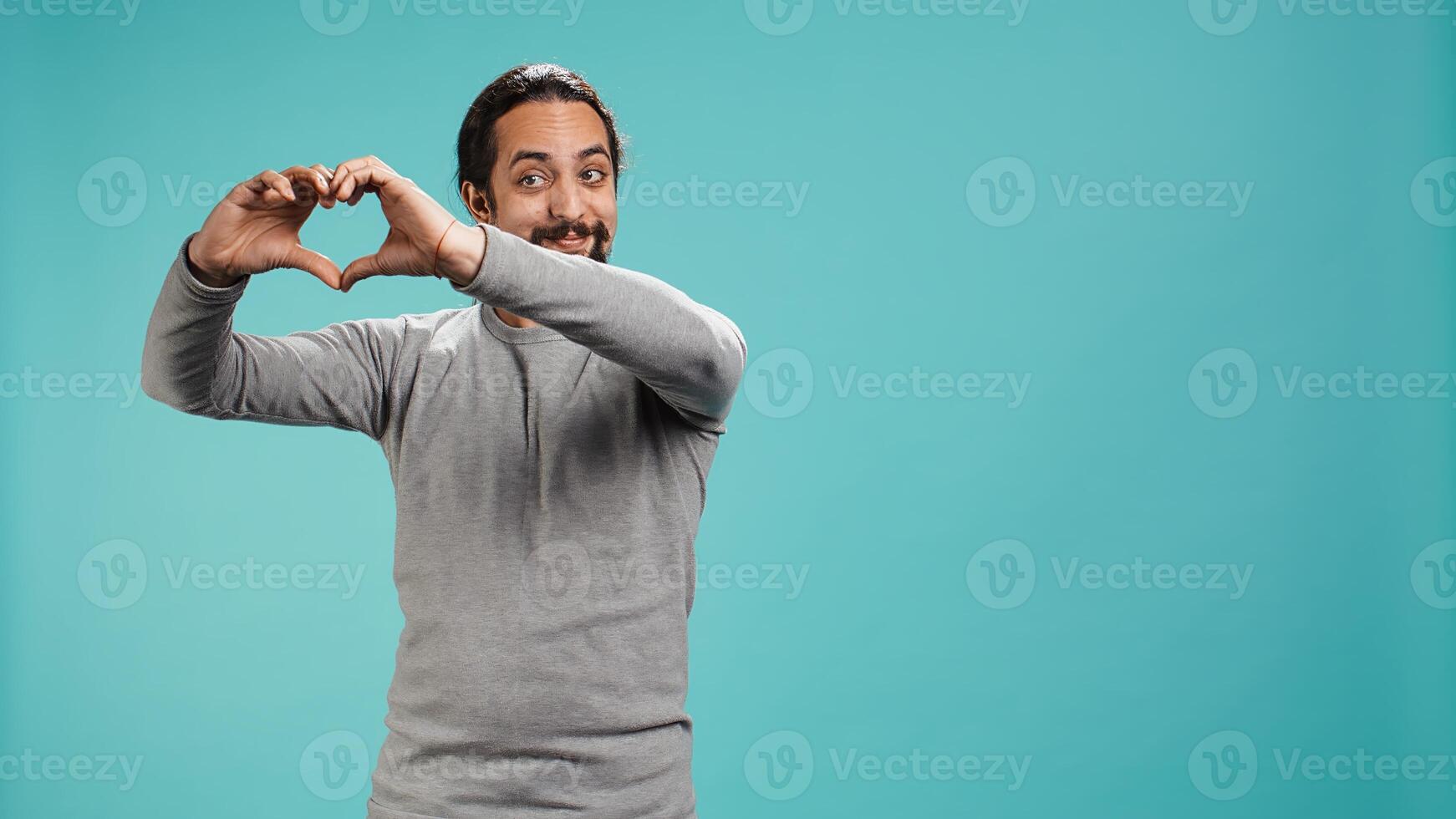 Portrait of joyful friendly man doing heart symbol shape gesture with hands. Smiling nurturing person showing love gesturing, isolated over blue studio background, camera B photo