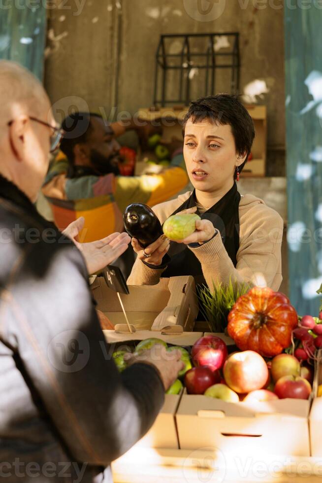 Woman stall holder selling healthy natural fruits and vegetables to elderly male customer. Young farmer displaying eco friendly seasonal products to consumer for sustainable living. photo