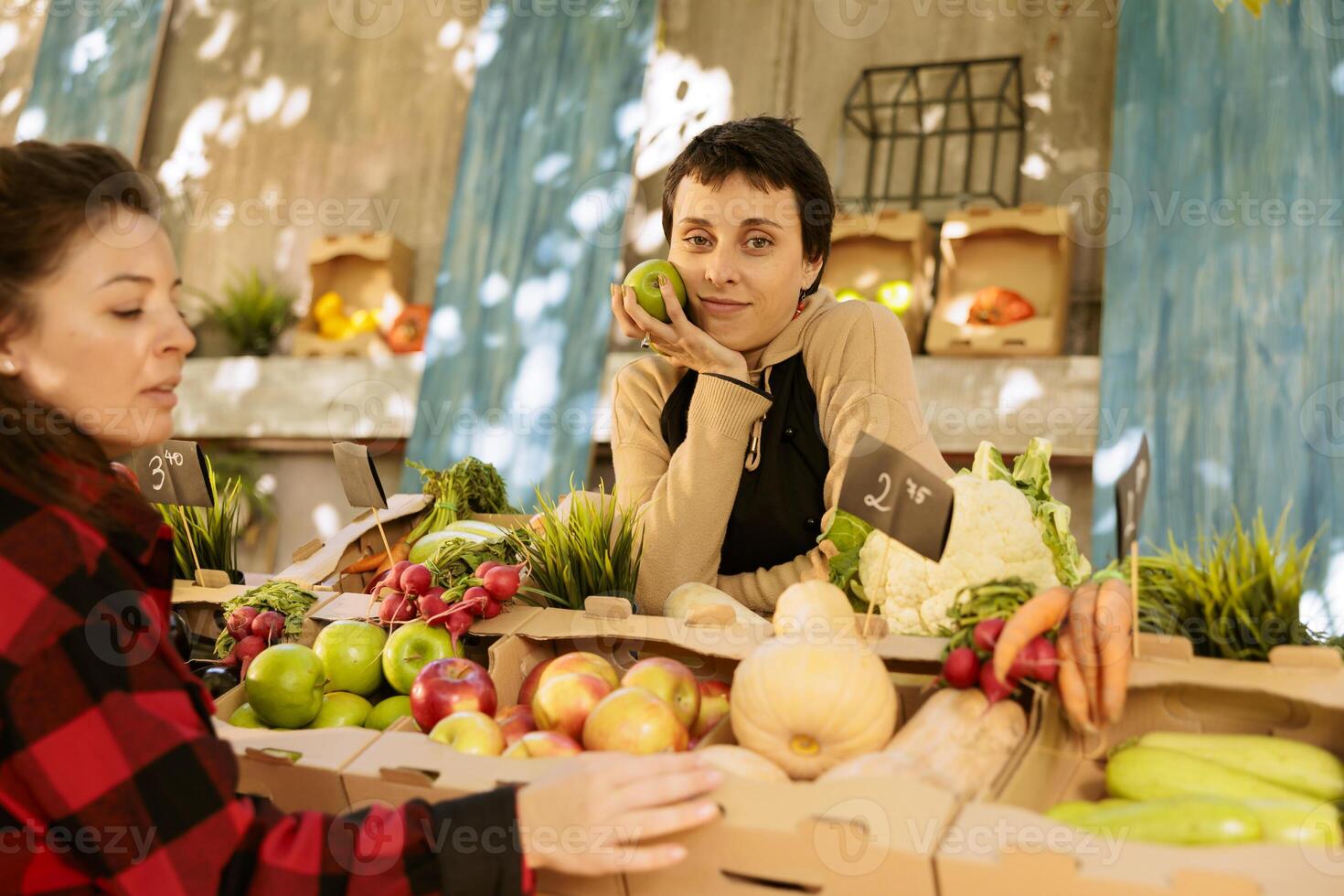 Female customer selecting fruits and vegetables while vendor stands behind counter with an apple in hand. Portrait of woman farmer selling variety of seasonal organic fresh produce at harvest fair. photo