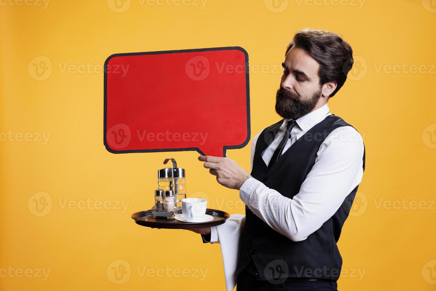 Professional butler holds speech bubble on camera, presenting isolated red empty billboard to create advertisement in studio. Young man waiter shows blank copyspace cardboard sign for ad. photo