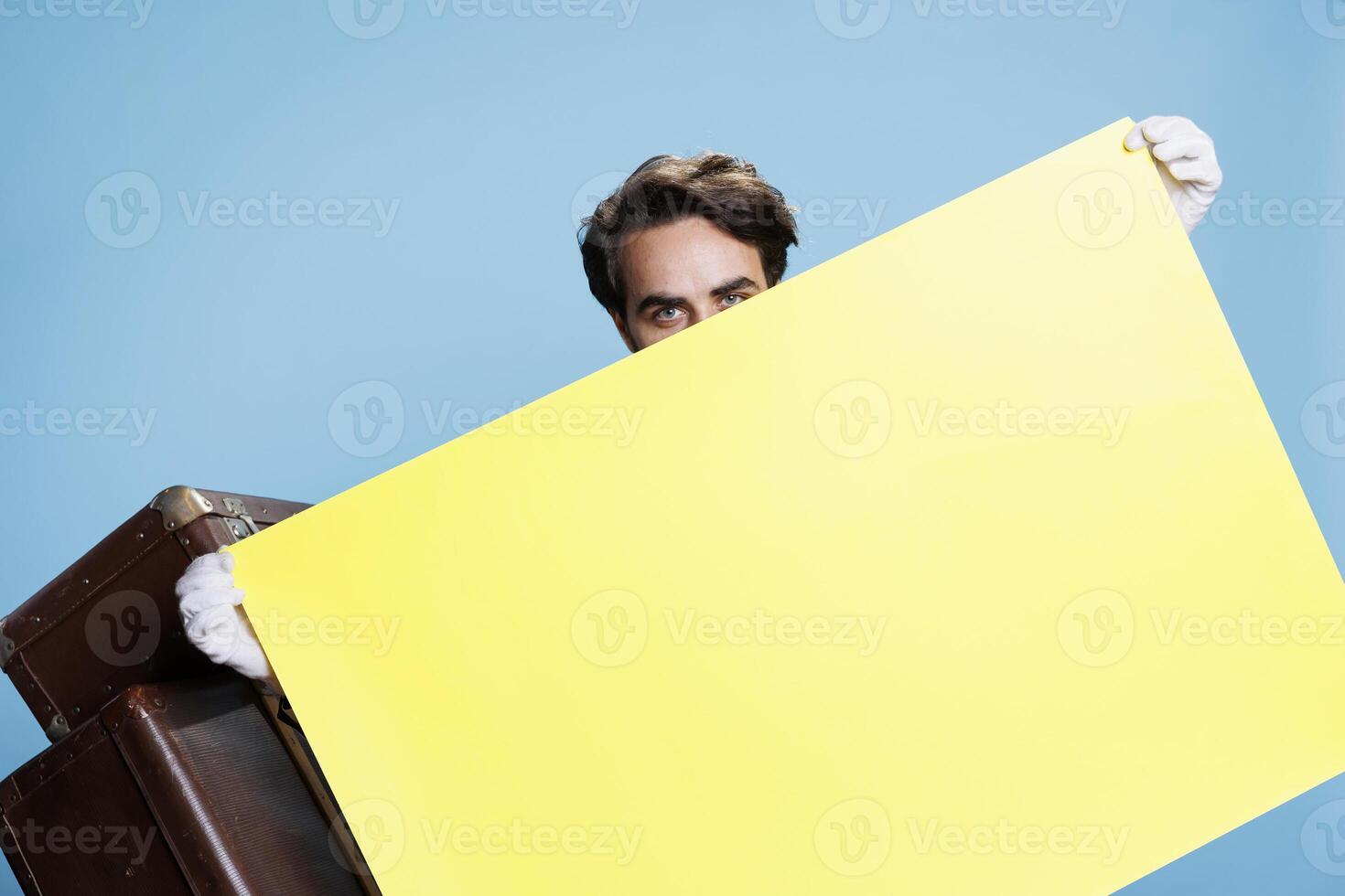 Hotel employee poses with blank banner on camera, showing isolated poster with empty copyspace for advertisement. Doorman in classy suit holds billboard ad sign over background. photo