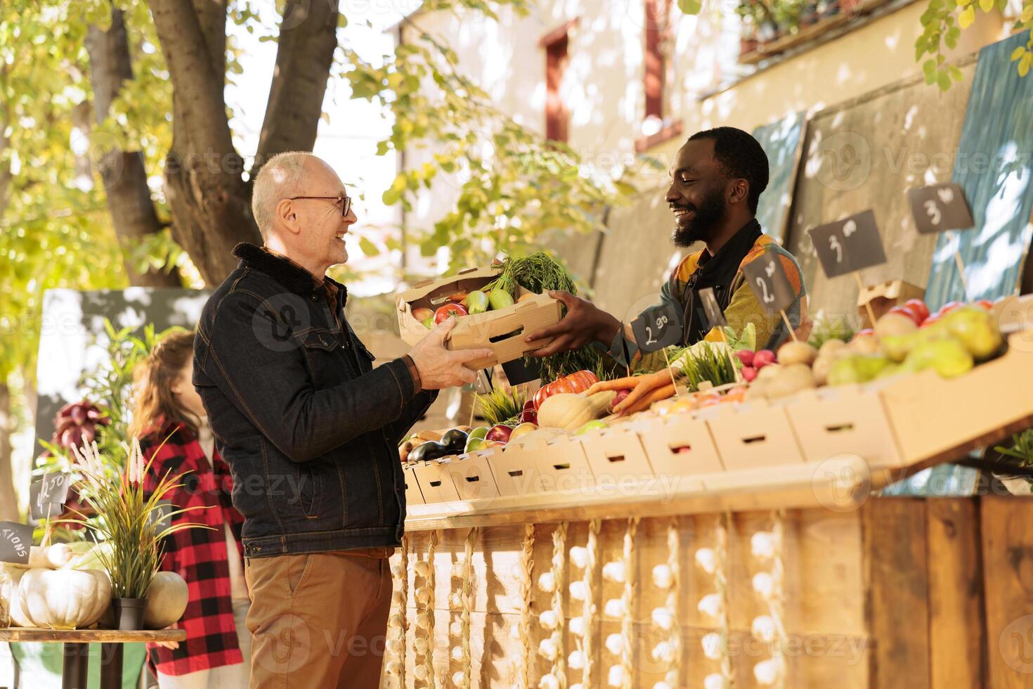 Happy elderly male person getting box of locally grown fruits and veggies at farmers market, visiting stand on sunny day. Smiling black man small business owner giving fresh organic produce. photo