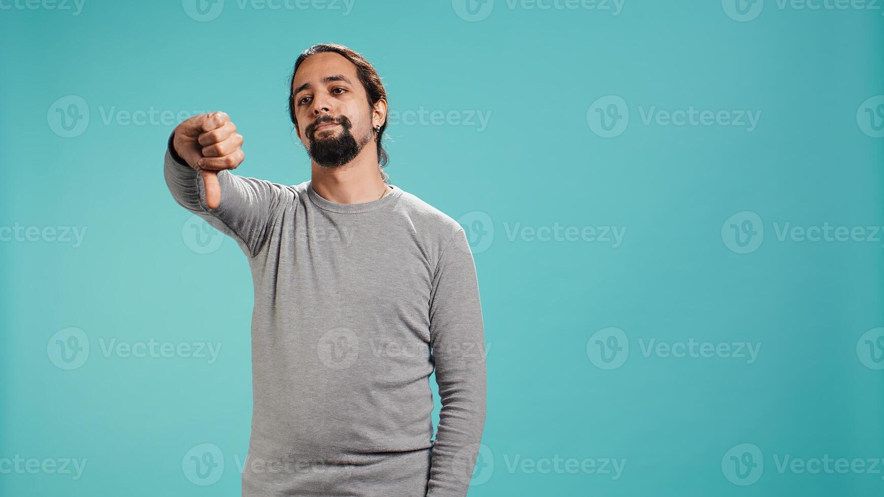 Upset man showing thumbs down sign gesturing, being disapproving. Disappointed Arab person against gaming, doing rejection hand gesture, isolated over studio background, camera B photo