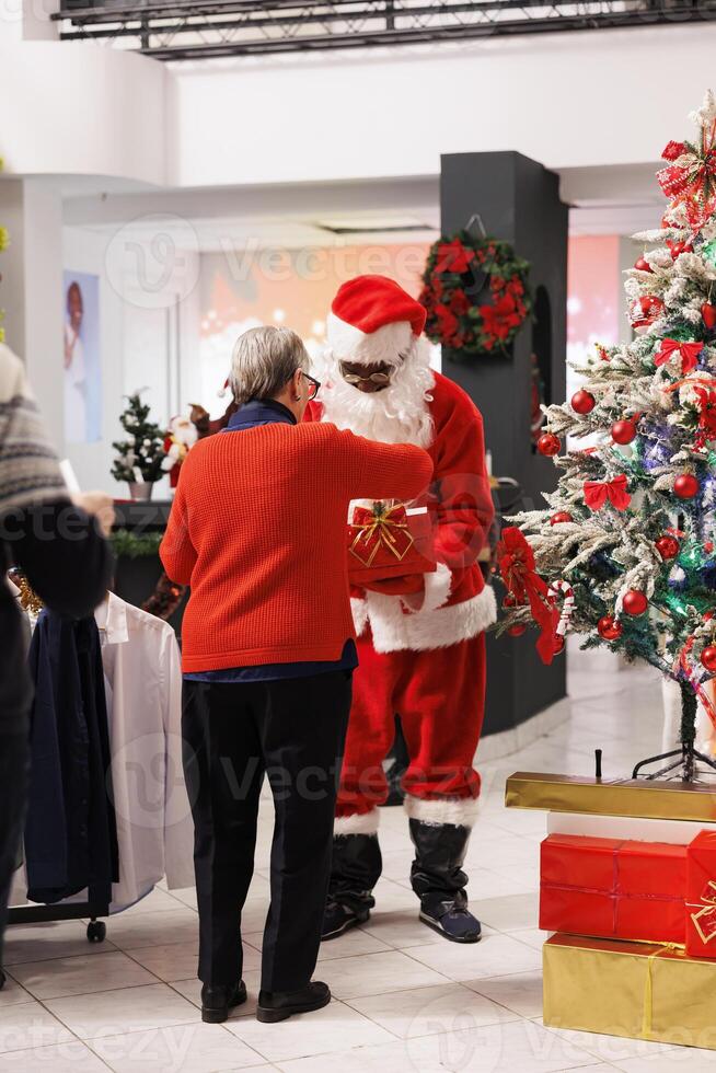 Visitors enter holiday prize draws, worker disguised as Santa Claus inserts cards with names written inside box. Older people waiting in festive store for contest results to win free attire. photo