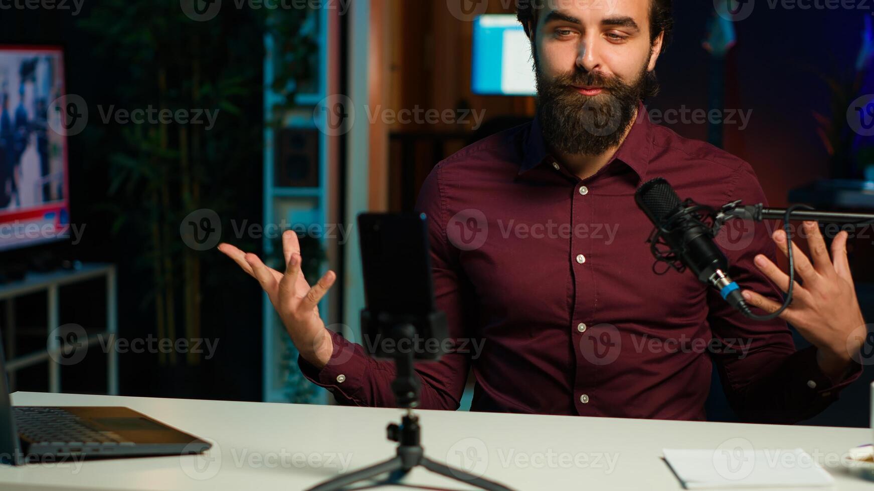 Influencer talks in front on smartphone attached to tripod and high quality mic in studio, discussing with viewers. Man using mobile phone and professional gear to record social media , camera A photo