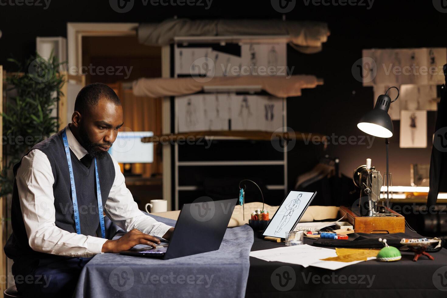 African american suitmaker looking online for outfit inspiration, comparing results with sketch drawings of sartorial garments. Senior couturier ordering sewing materials on laptop in tailoring studio photo