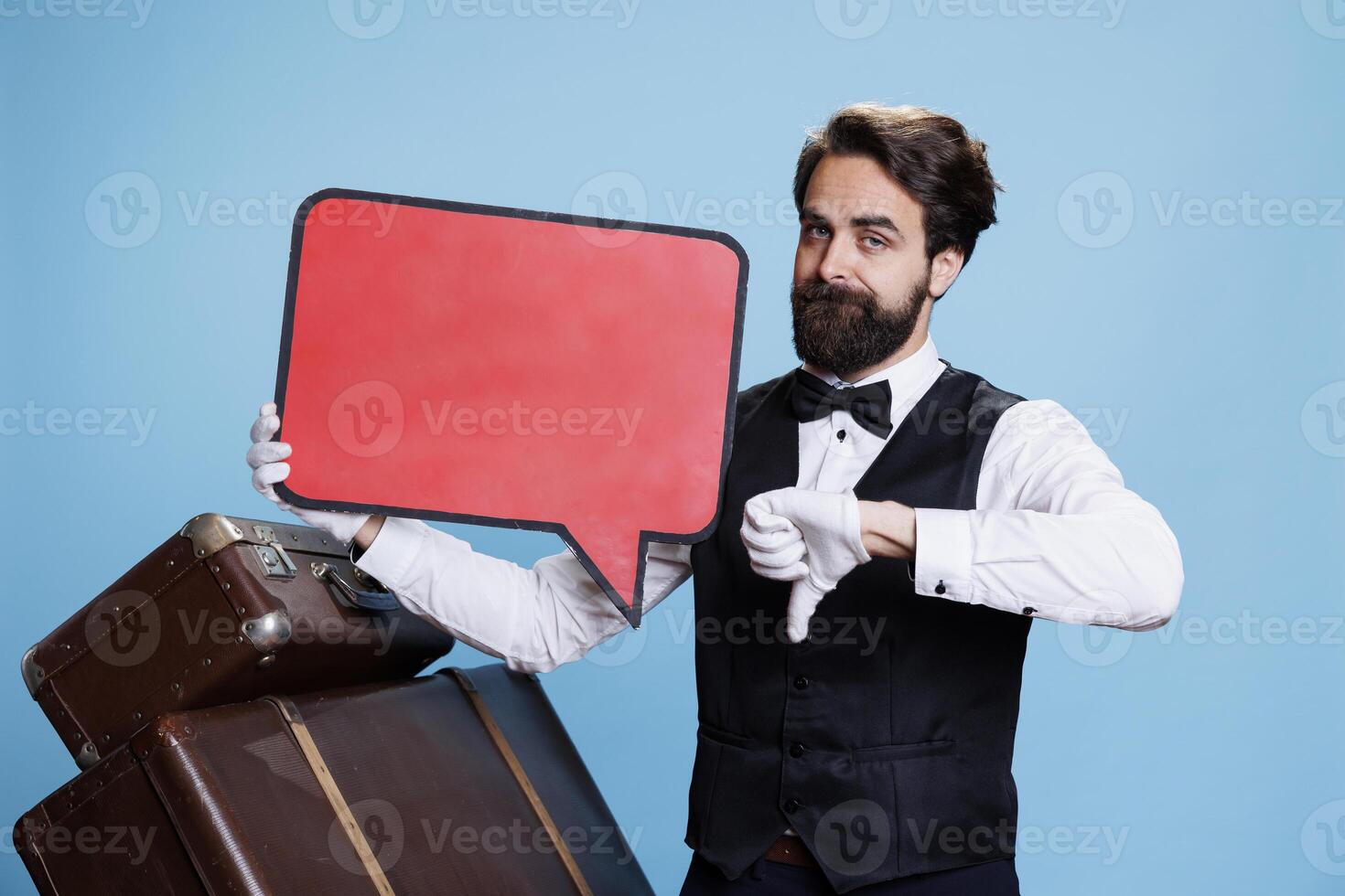 Stylish doorman holds red speech bubble and giving thumbs down in blue studio, creating marketing ad on empty cardboard sign. Hotel porter using empty isolated board, feeling displeased. photo