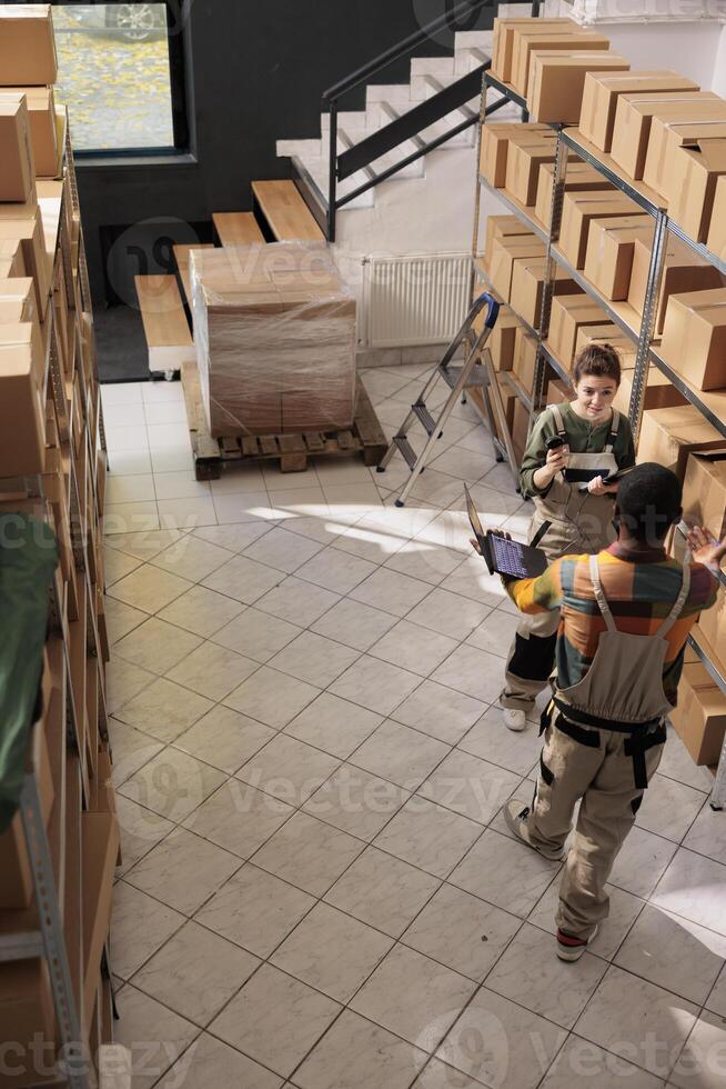 Diverse team discussing about invoice code on tablet trying to find customer parcel in storage room. Warehouse employee wearing protective overall while preparing orders for delivery. Aerial view photo