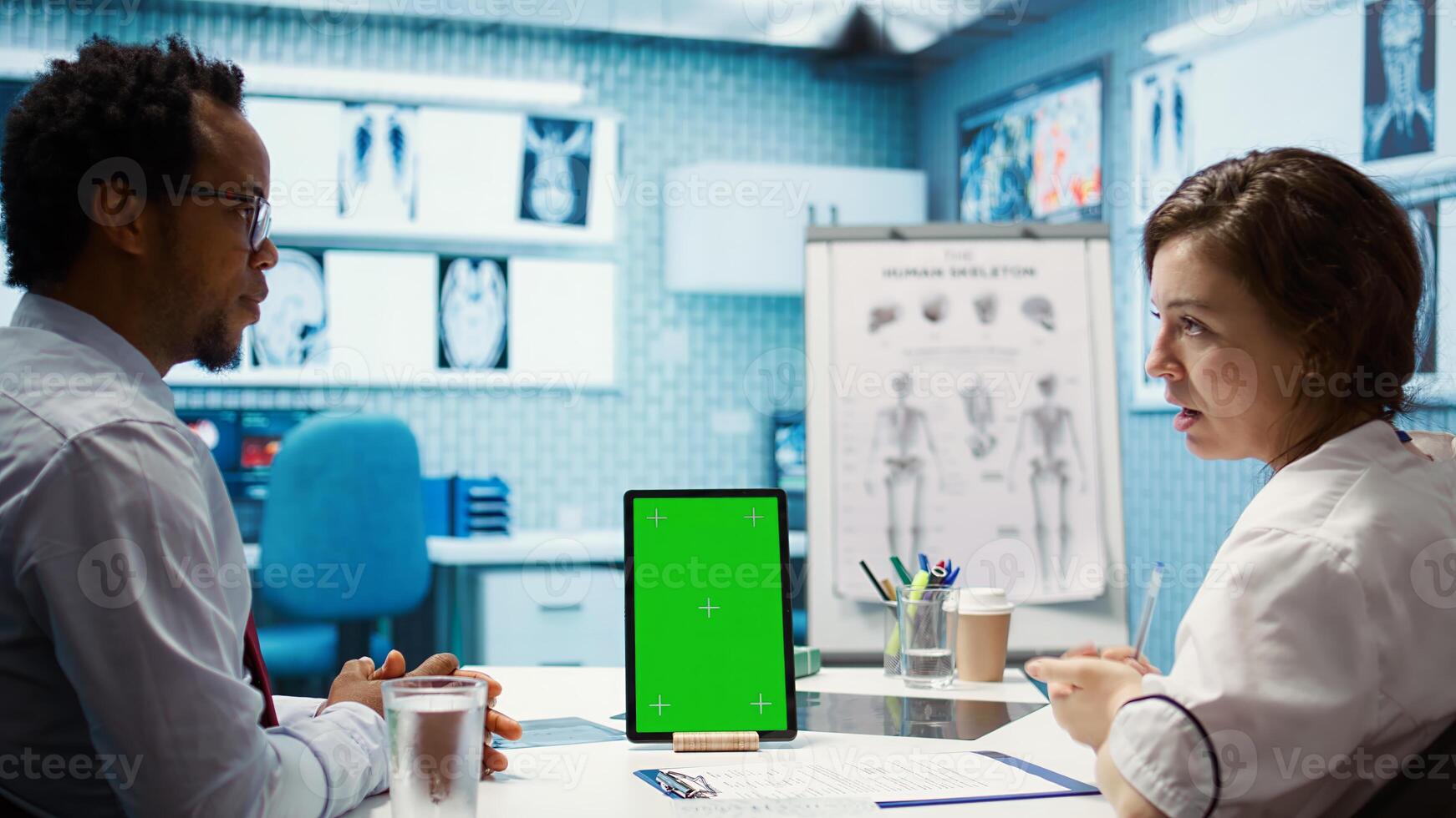 General practitioner pointing at green screen mockup on device, discussing with her patient about recovery treatment. Doctor doing checkup visit on insurance with a man. Camera B. photo