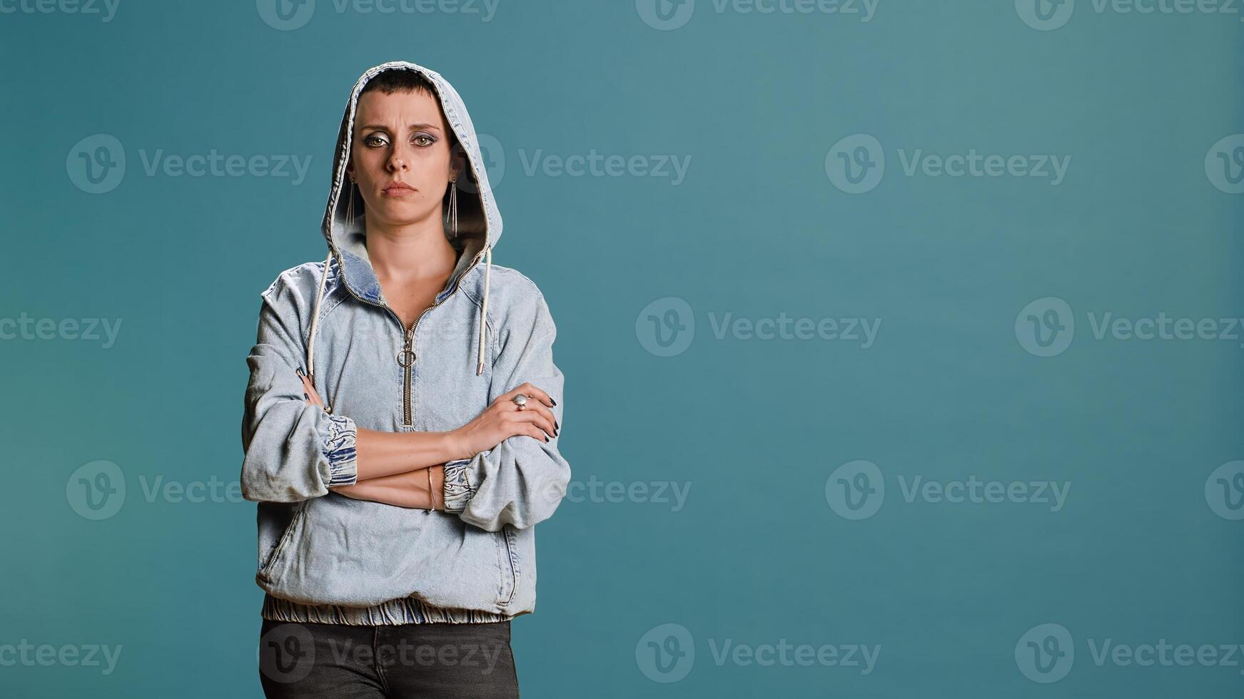 Bold serious woman putting her hood on and acting tough in studio, posing with arms crossed and showing confidence in her street style. Modern independent person acts as a gangster. Camera B. photo