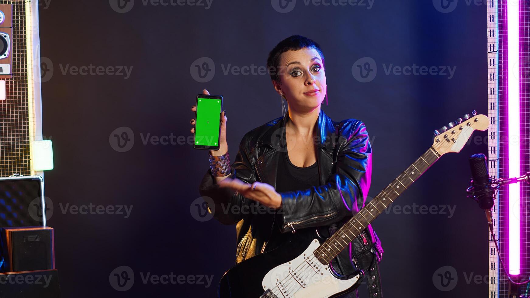 Woman rock musician playing guitar next to green screen display on device, doing a live music performance on stage at the nightclub. Artist rocker acting cool and rebellious with edgy style. Camera A. photo