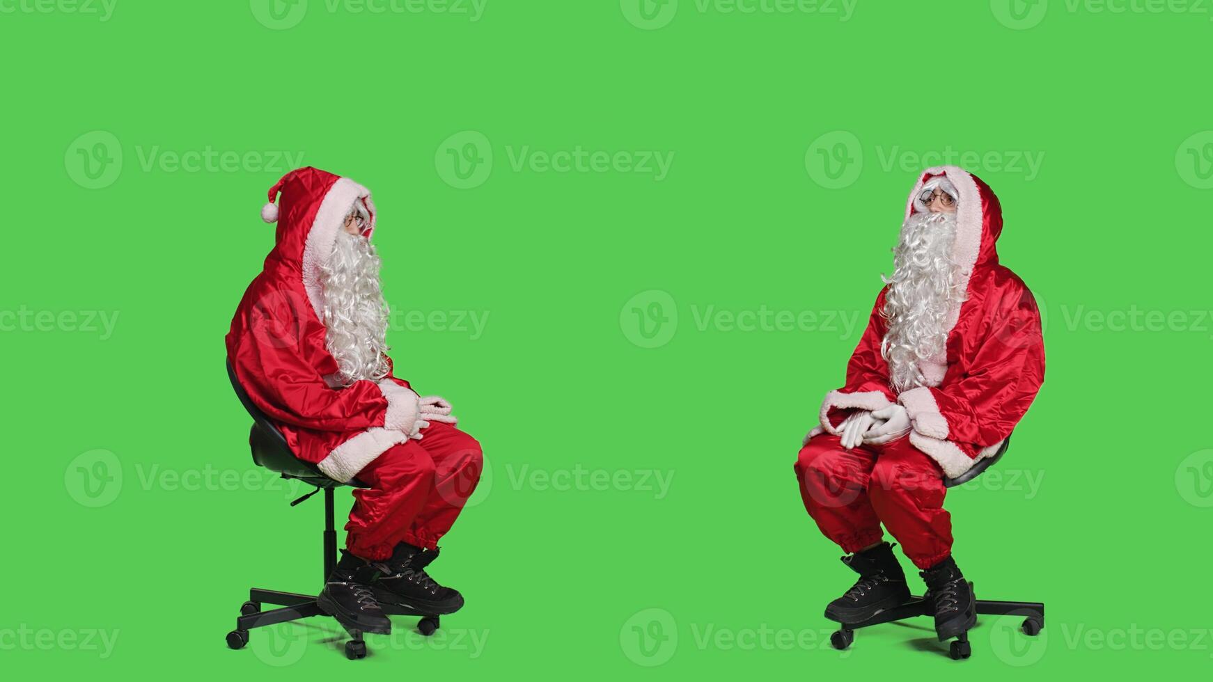 Santa claus embodiment poses on chair, sitting and looking around while he wears festive iconic red costume with white beard. Saint nick famous winter character on full body greenscreen. photo