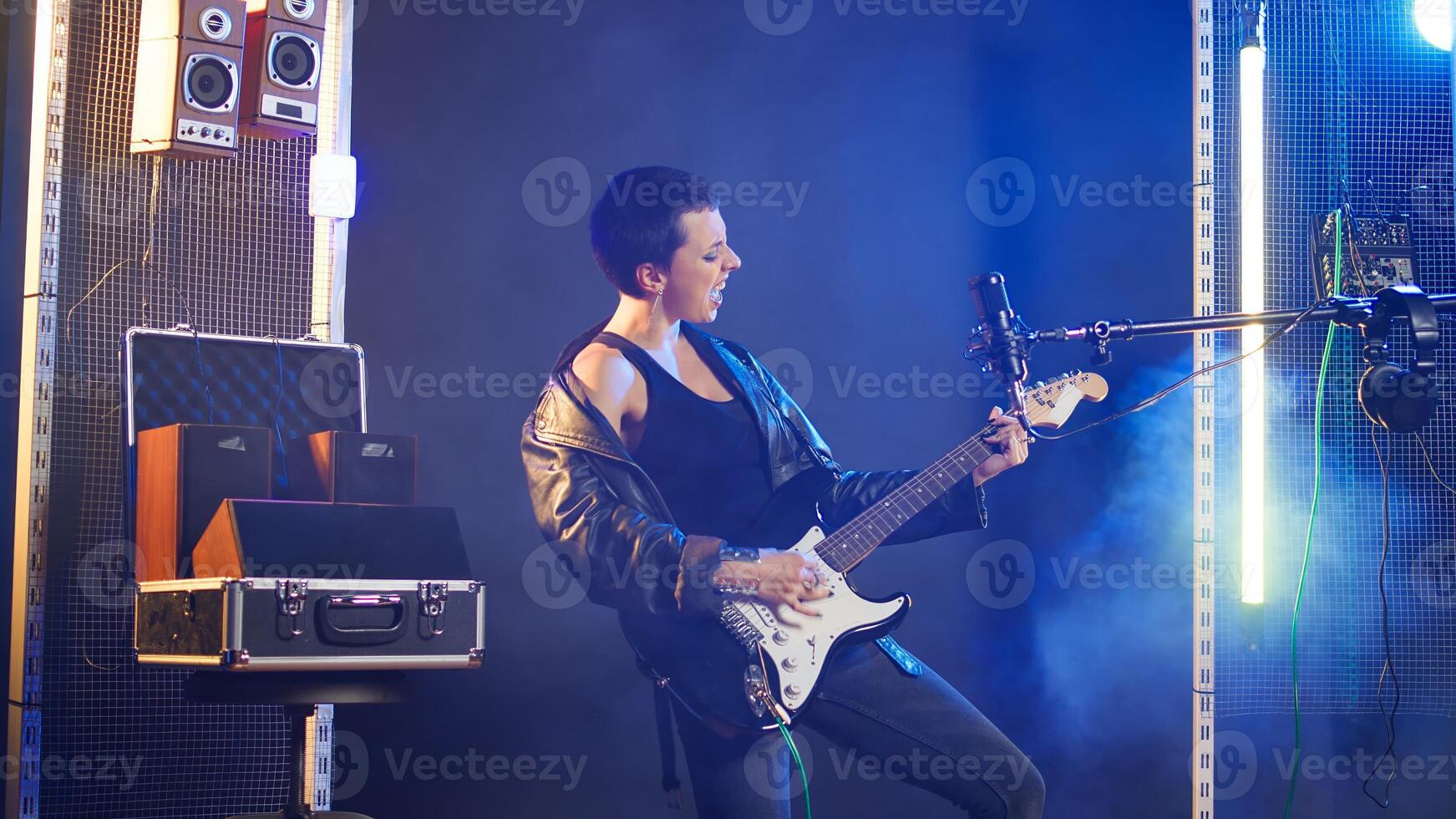 Rebellious edgy woman playing guitar in a modern studio setting, performing an iconic and bold song. Independent energetic artist singing with confidence at microphone, music lover. Camera A. photo