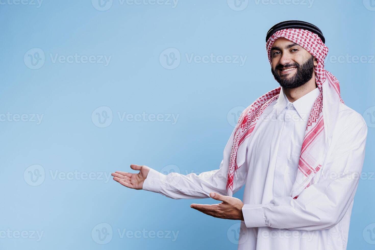 Arab man in thobe and ghutra pointing to side with hands, advertising product studio portrait. Muslim person wearing traditional clothes showing to right direction with arms photo