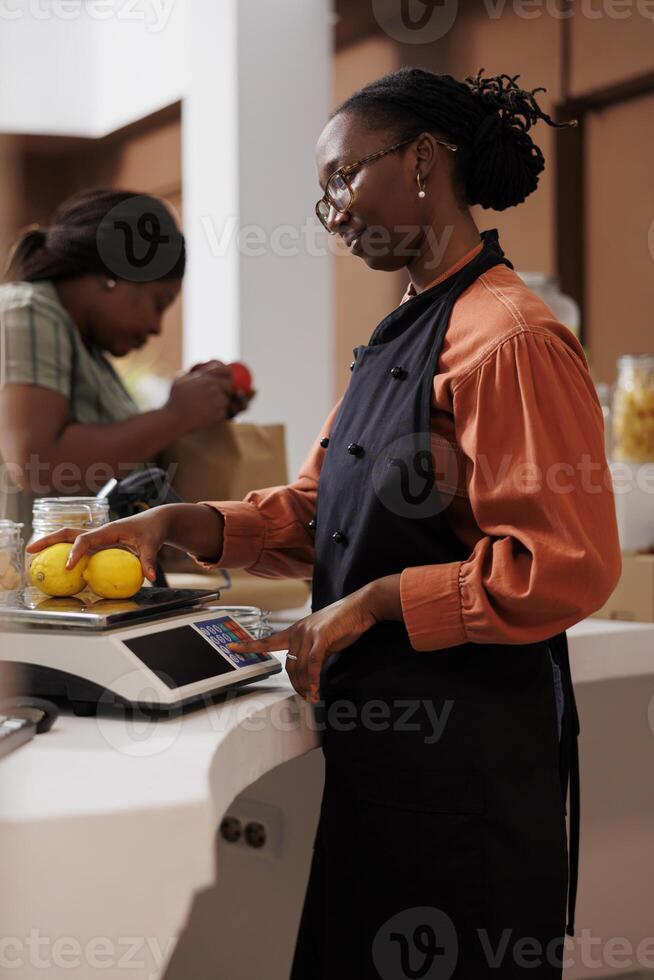 Female farmer vendor in zero waste shop at checkout counter processing customer order. Young client buying organic nutritious food from her ecofriendly local neighborhood store. photo