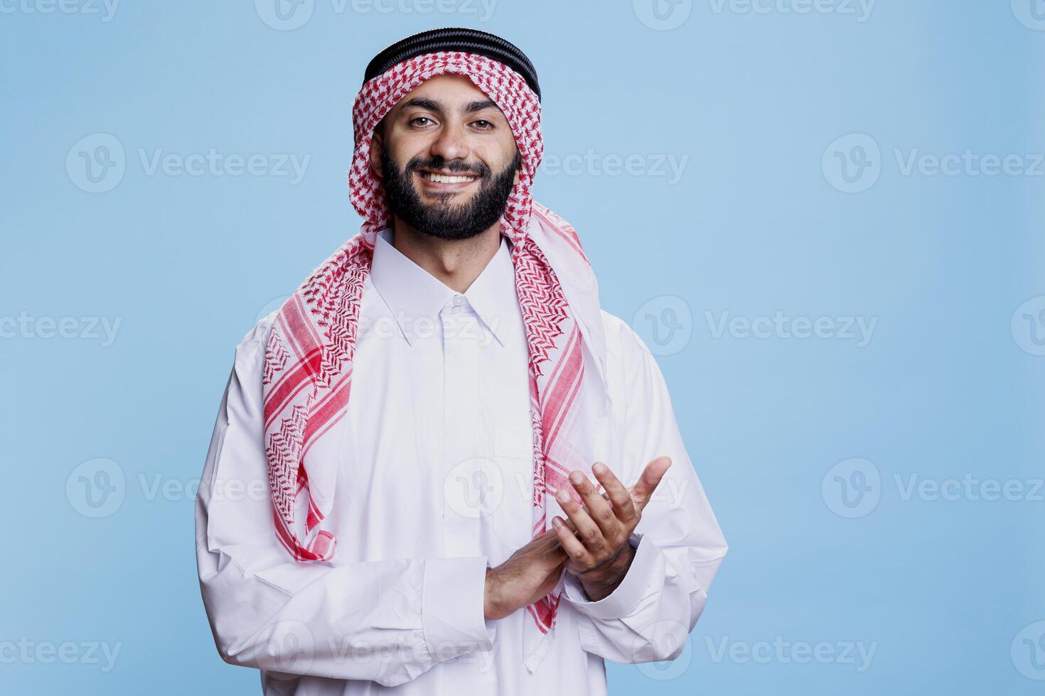 Smiling muslim man showing applause gesture and looking at camera with excited expression. Cheerful arab wearing islamic thobe and ghutra applauding at performance studio portrait photo