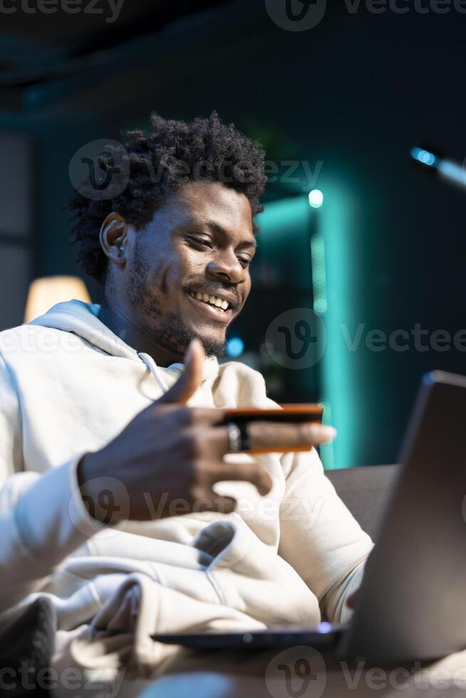 BIPOC man having fun, adding payment method on website while in apartment living room. Joyous person delighted to do online shopping, carefully typing credit card information photo