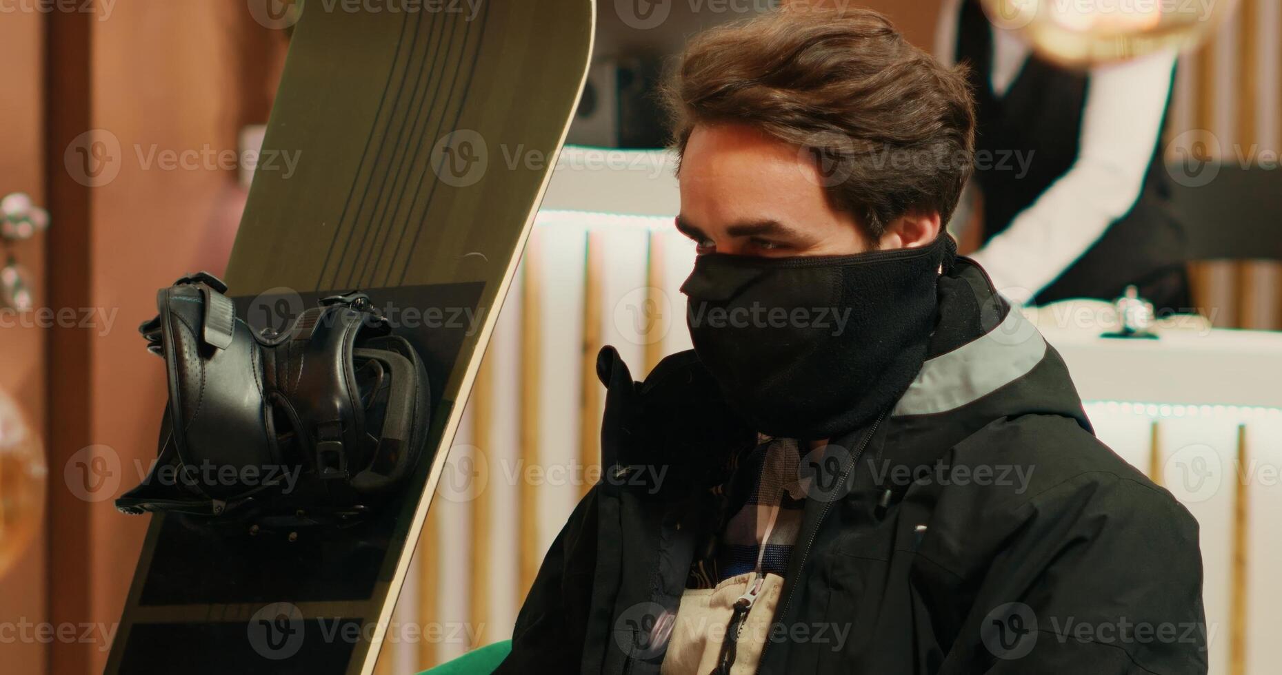 Young man in winter sport attire preparing to face the cold weather and go down snowy slopes with snowboard. Person sitting in lounge area at exclusive alpine resort waiting for check in. photo