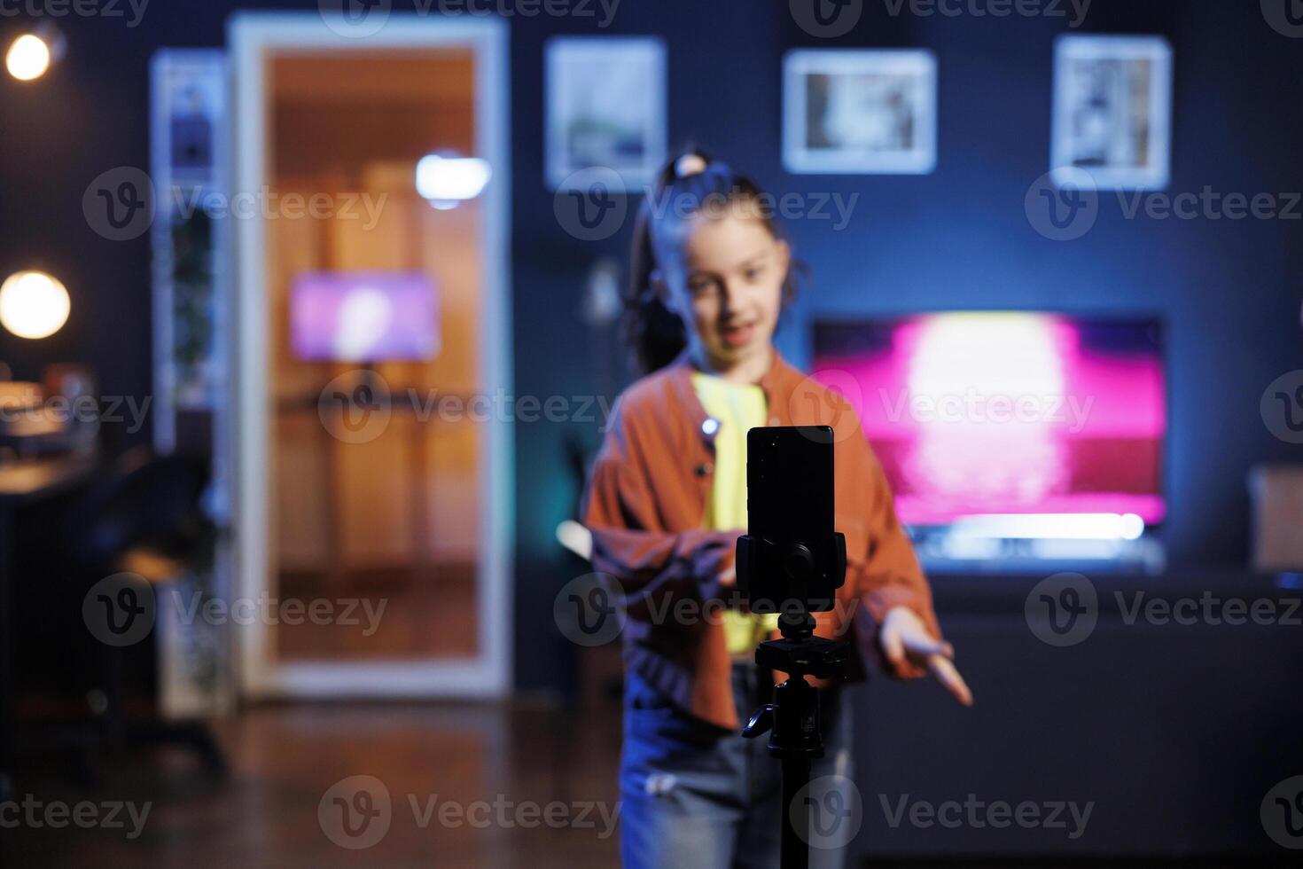 Focus shot on cellphone on tripod used by girl dancing and singing, shooting for social media. Kid in blurry background entertains subscribers with professional choreography recorded with phone photo