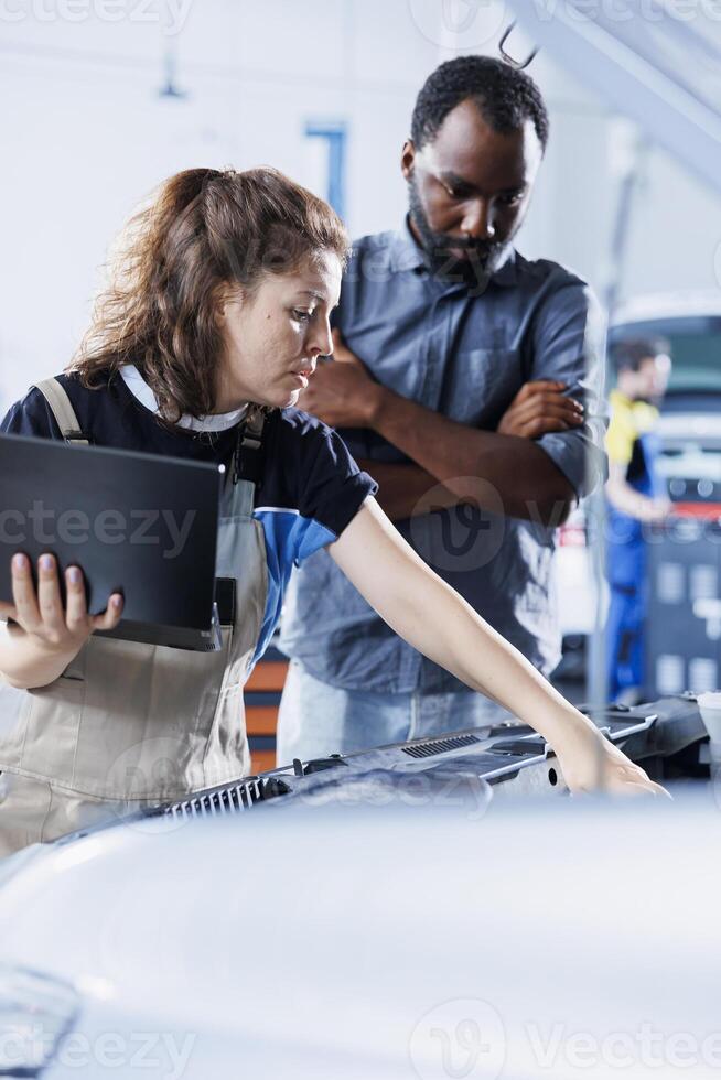 Friendly mechanic at auto repair shop conducts annual vehicle checkup, informing client about needed engine replacement. Automotive expert talks with customer after finishing car inspection photo