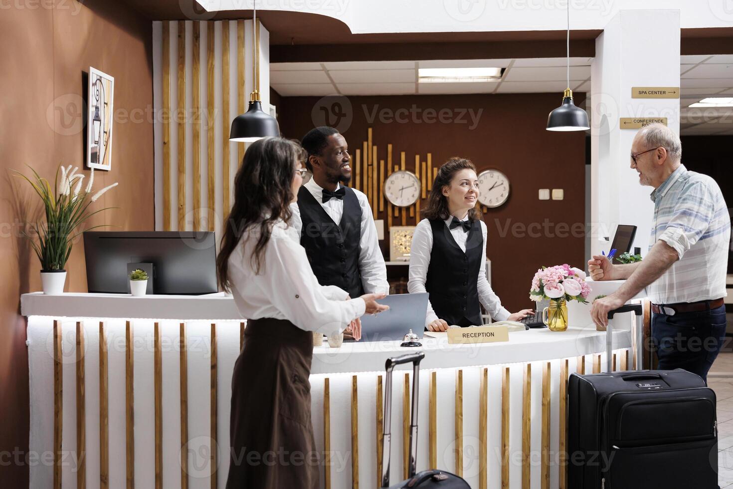Retired senior couple arrives at luxurious resort for vacation, assisted by friendly employees at reception desk. Hotel lobby is elegant with lounge area. Smooth check-in process for elderly tourists. photo