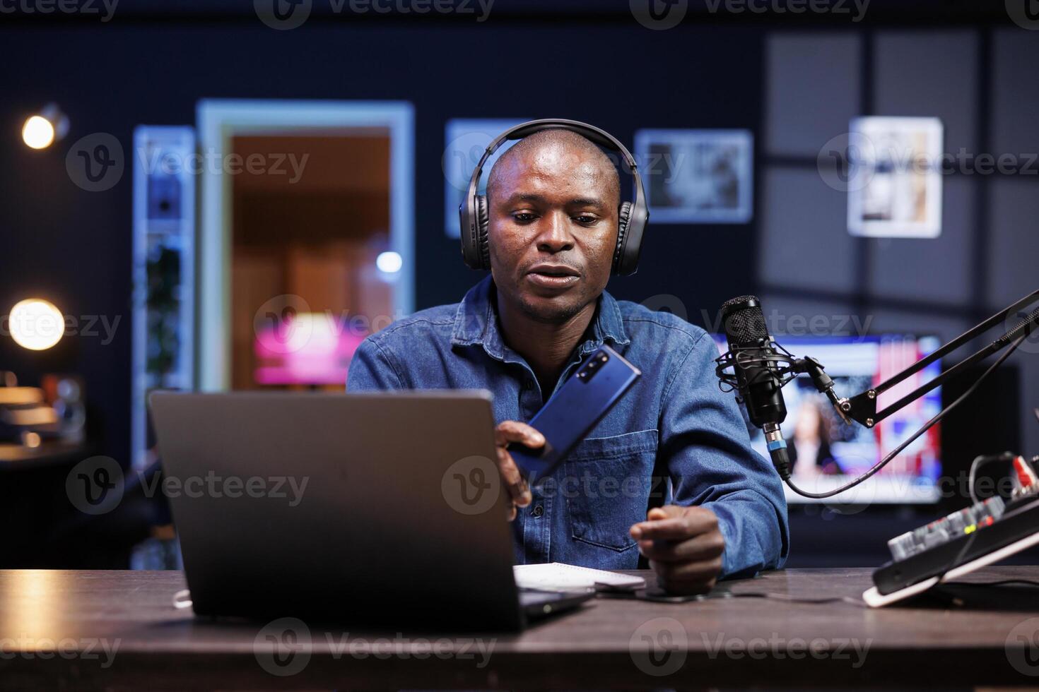 African American man in portrait holding a smartphone and showcasing it on a live feed at home. Black male influencer reviewing new mobile device on the internet, filming with recording equipment. photo