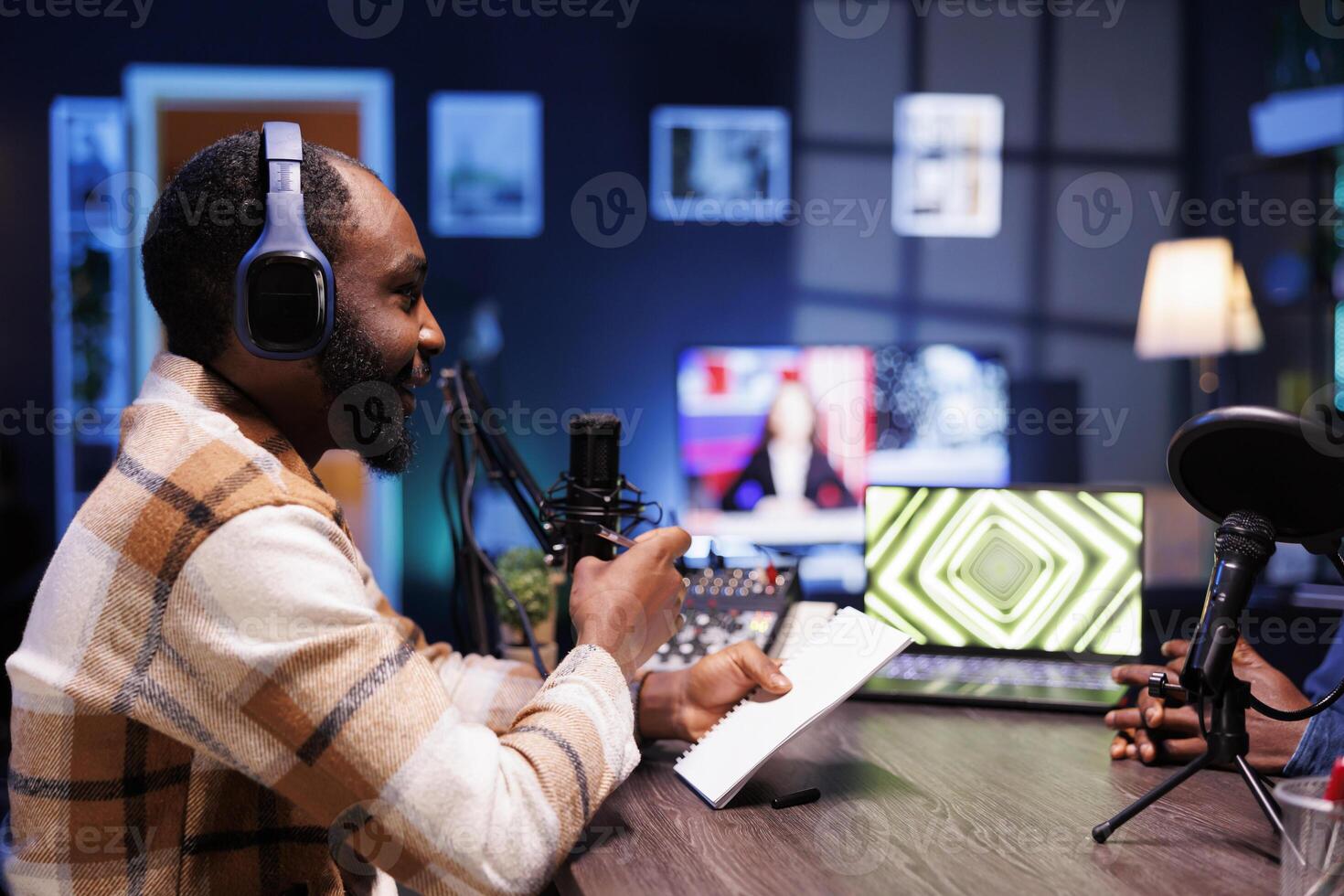 Male talk show host holding a notebook and interviewing a guest, having online discussion for their listeners. African american man using recording equipment to create a radio program at home. photo
