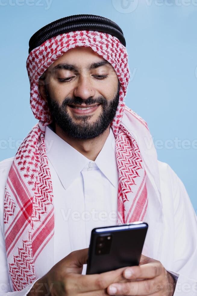 Smiling arab man wearing islamic traditional clothes typing message on smartphone. Muslim person dressed in thobe and ghutra chatting in social media using mobile phone with cheerful expression photo