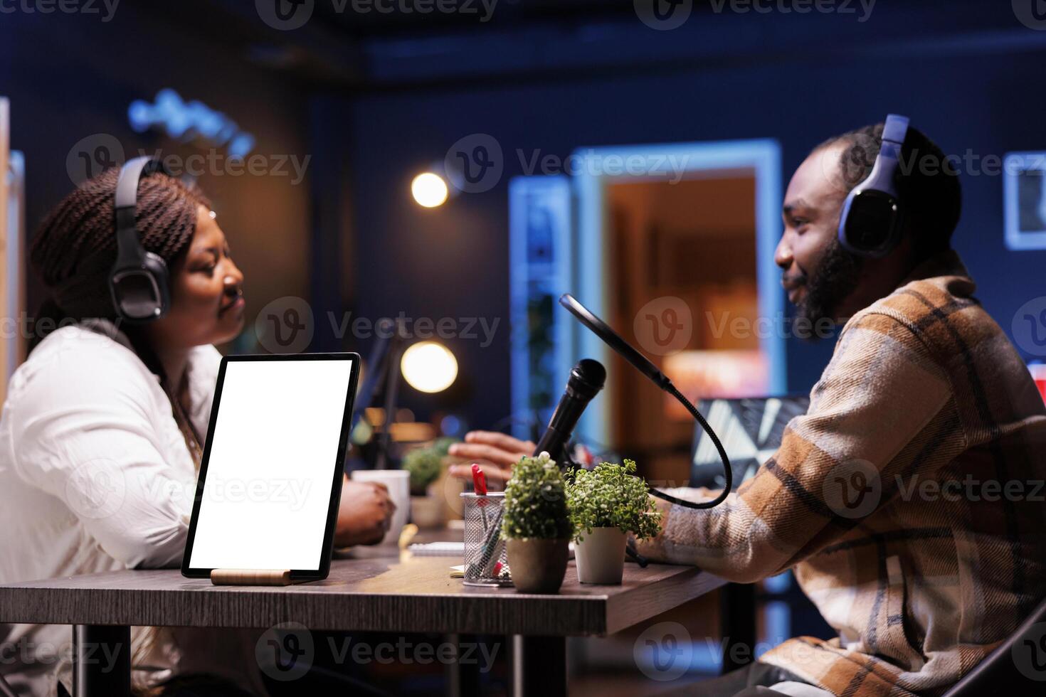Young content creators using headphones are broadcasting live while tablet shows blank white screen. Black people using podcast equipment while device positioned on desk displays chromakey template. photo