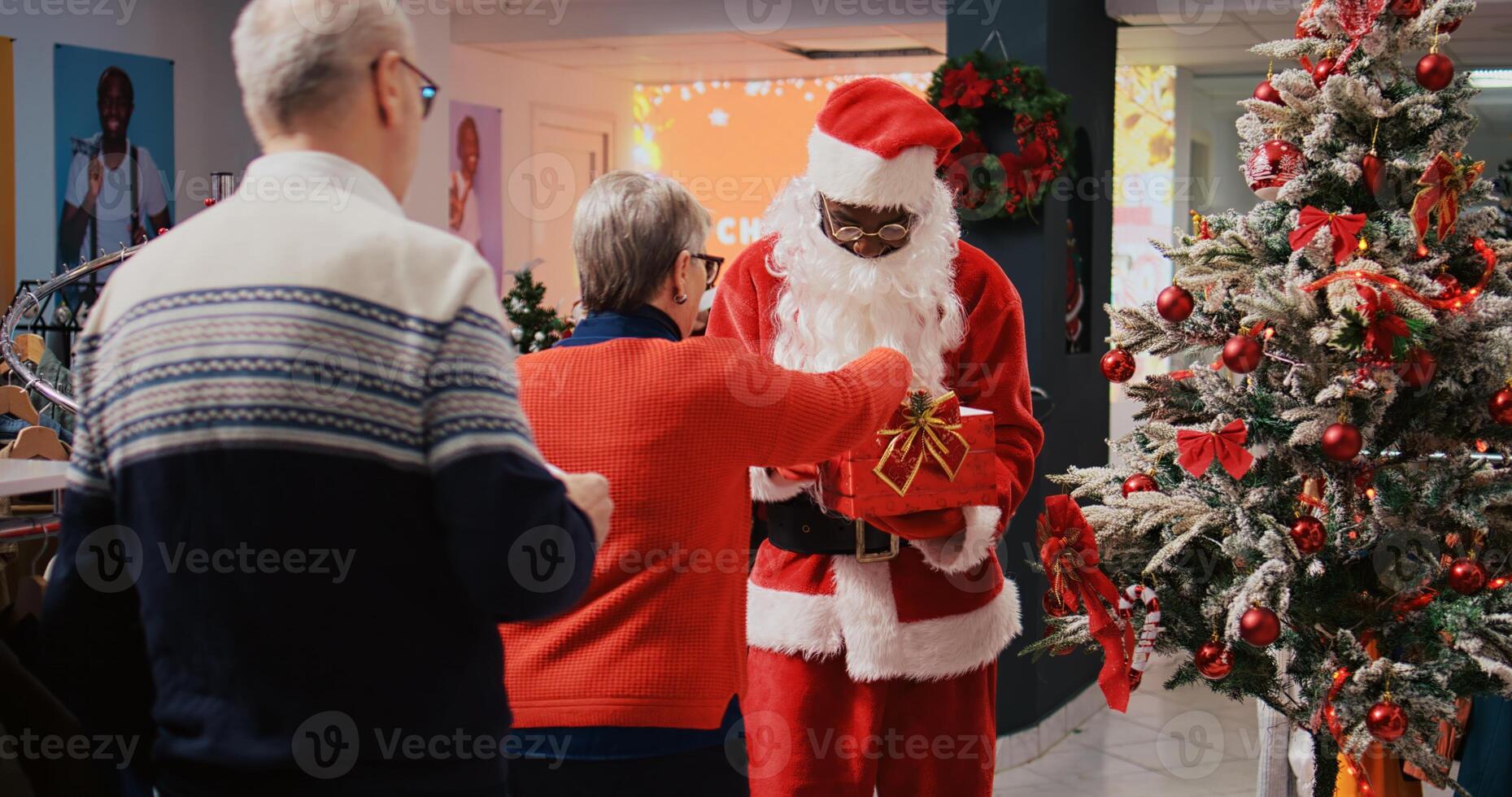 Employee wearing Santa Claus suit holding raffle contest in Christmas decorated shopping mall clothing shop during winter holiday season, inviting elderly couple to participate photo