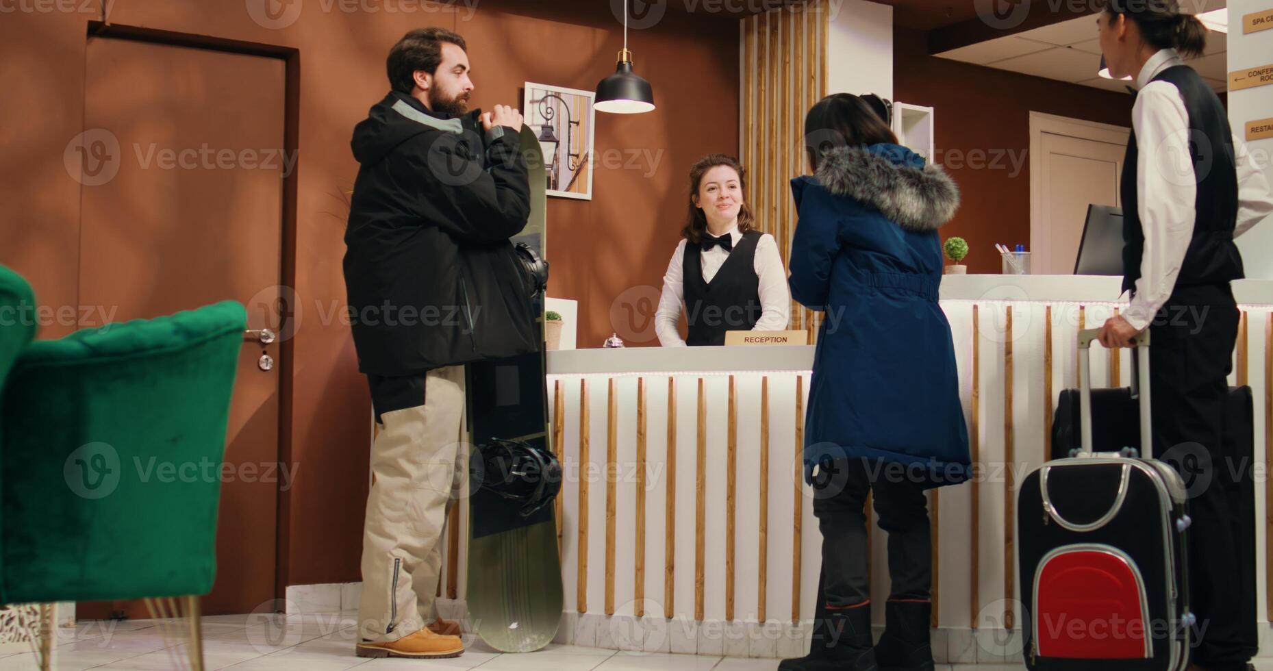 Active people enter alpine resort lobby, approaching reception staff to see room reservation while carrying skiing equipment. Young couple at mountain hotel preparing for winter sport activity. photo