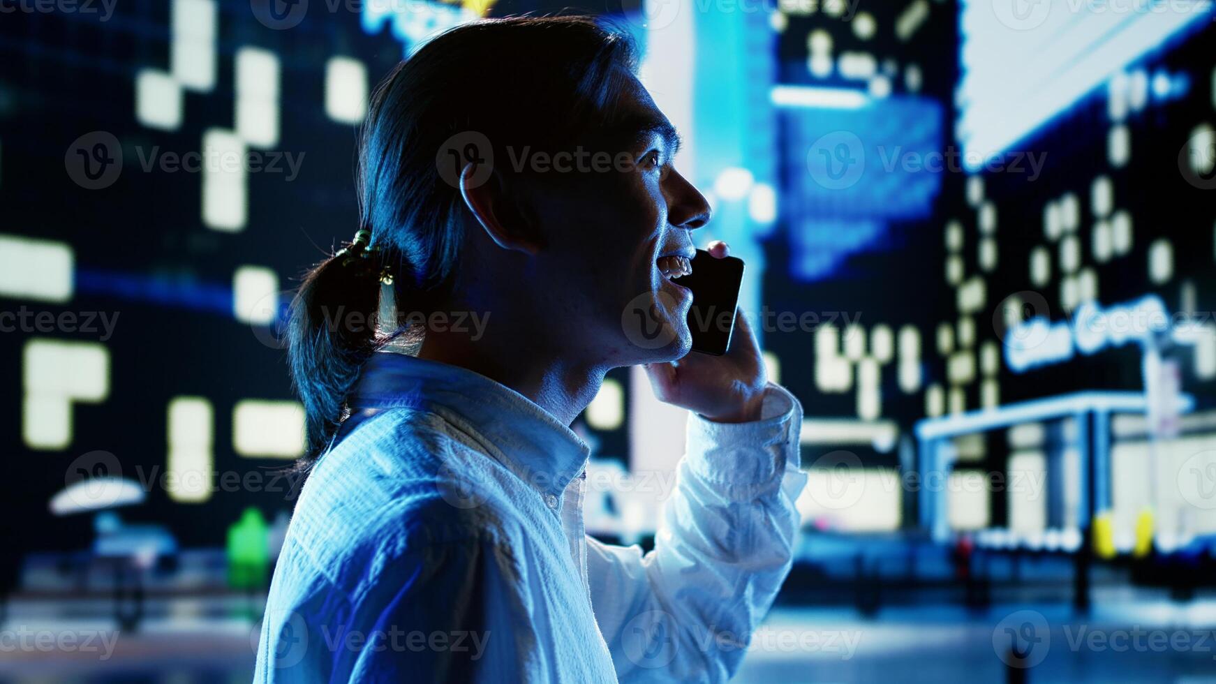Asian man wandering around city avenues during nighttime, talking over the phone with friend while looking upwards to surrounding buildings in metropolitan urban environment photo