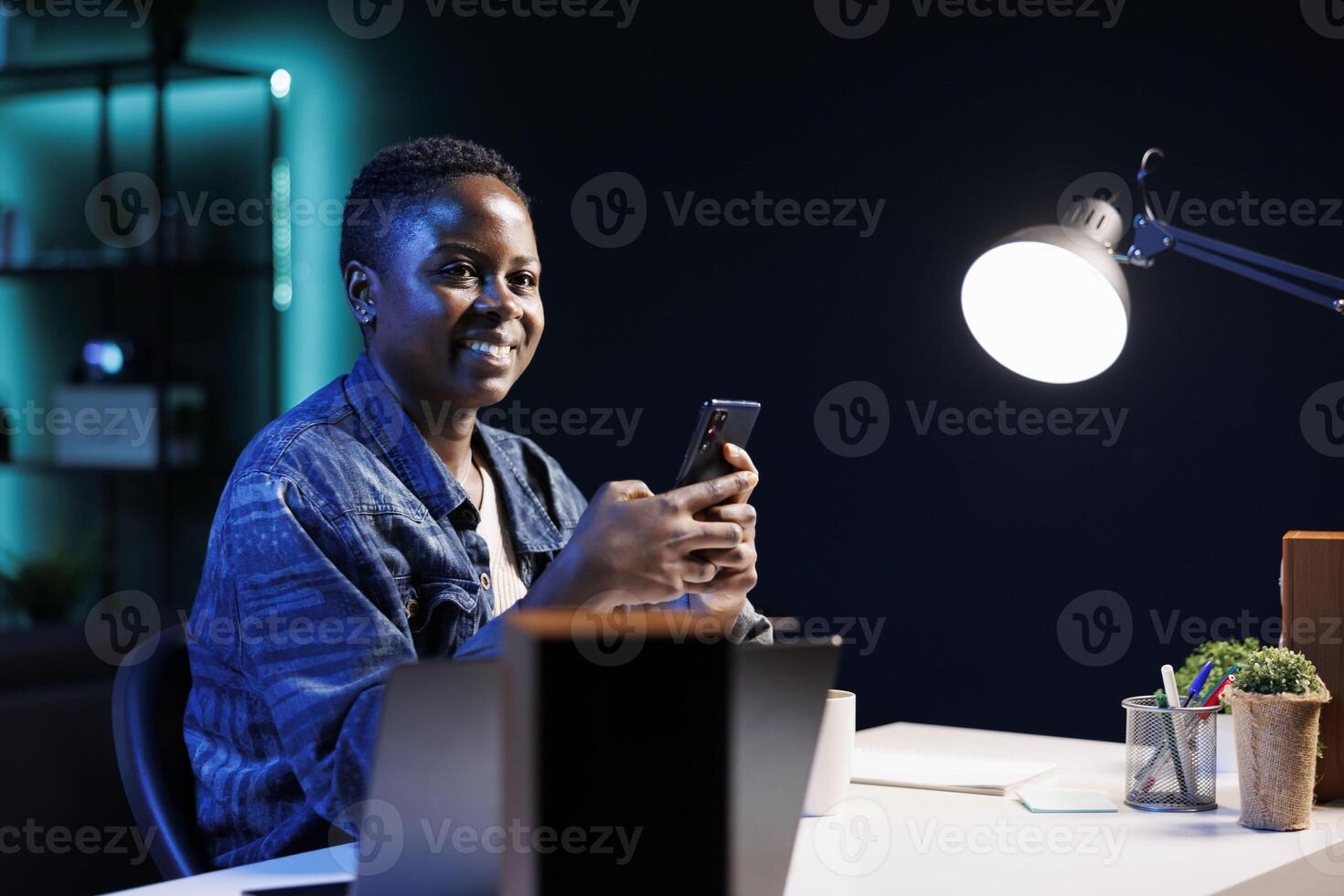Self employed African American lady grasping her smartphone for texting messages while working from home at table. Portrait of black woman chatting on social media with her electronic cell phone. photo
