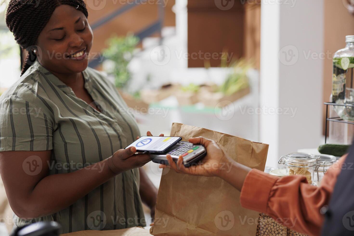 Cheerful black woman buying bio food with mobile payment at retail store checkout counter using credit card on her phone, while smiling and being served by a smiling vendor photo