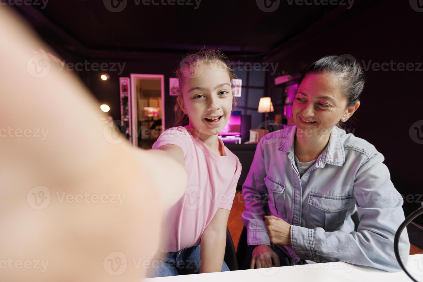 Happy girl next to her mother films with selfie camera in pink neon lit living room used as vlogging studio. Young media star captures footage with high tech camera, helped by parent, POV shot photo