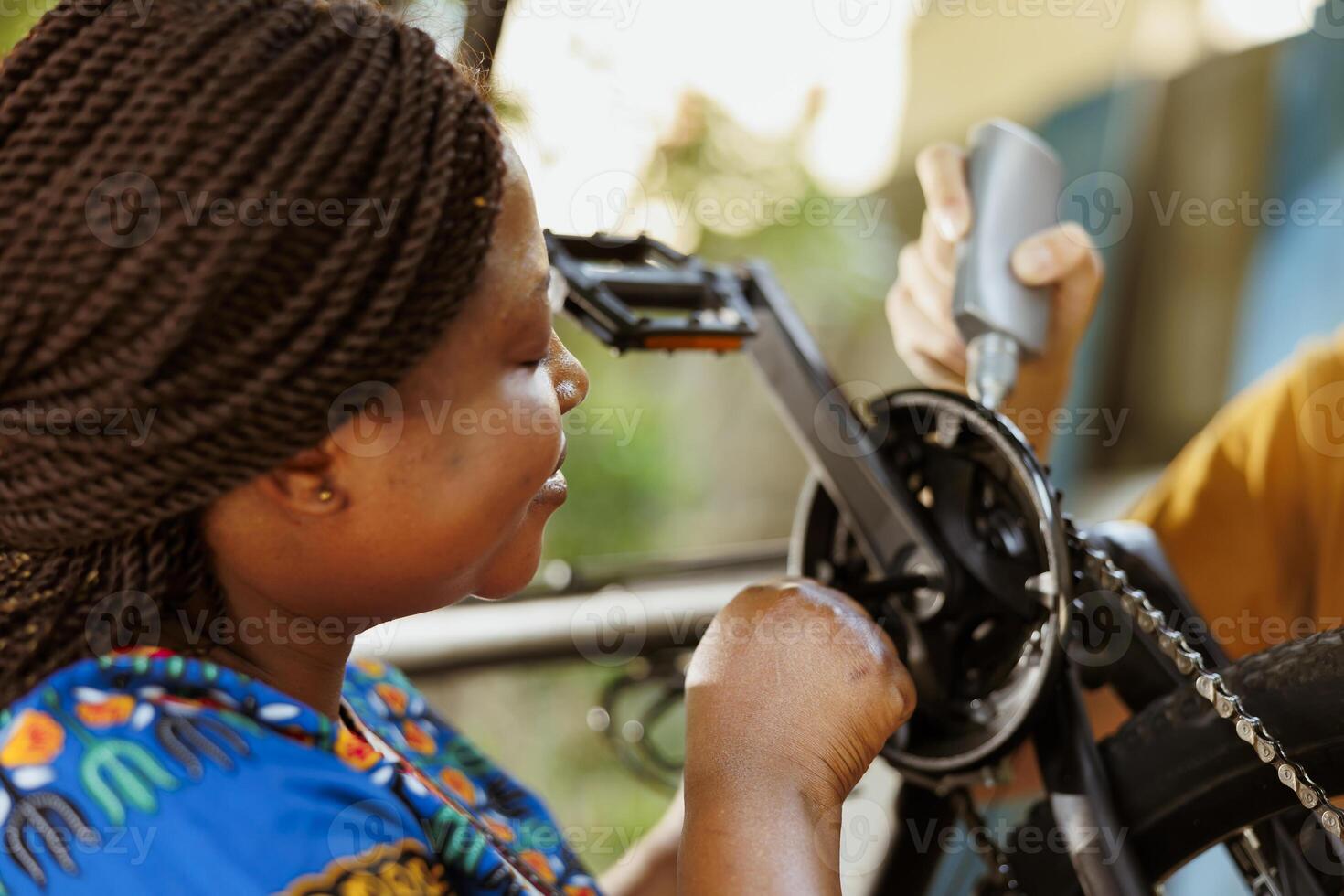Active and enthusiastic black woman doing annual bike maintenance by making adjustments to pedals and checking gears in home yard. Using specialized tools boyfriend and girlfriend lubricating and fastening bicycle parts outside. photo