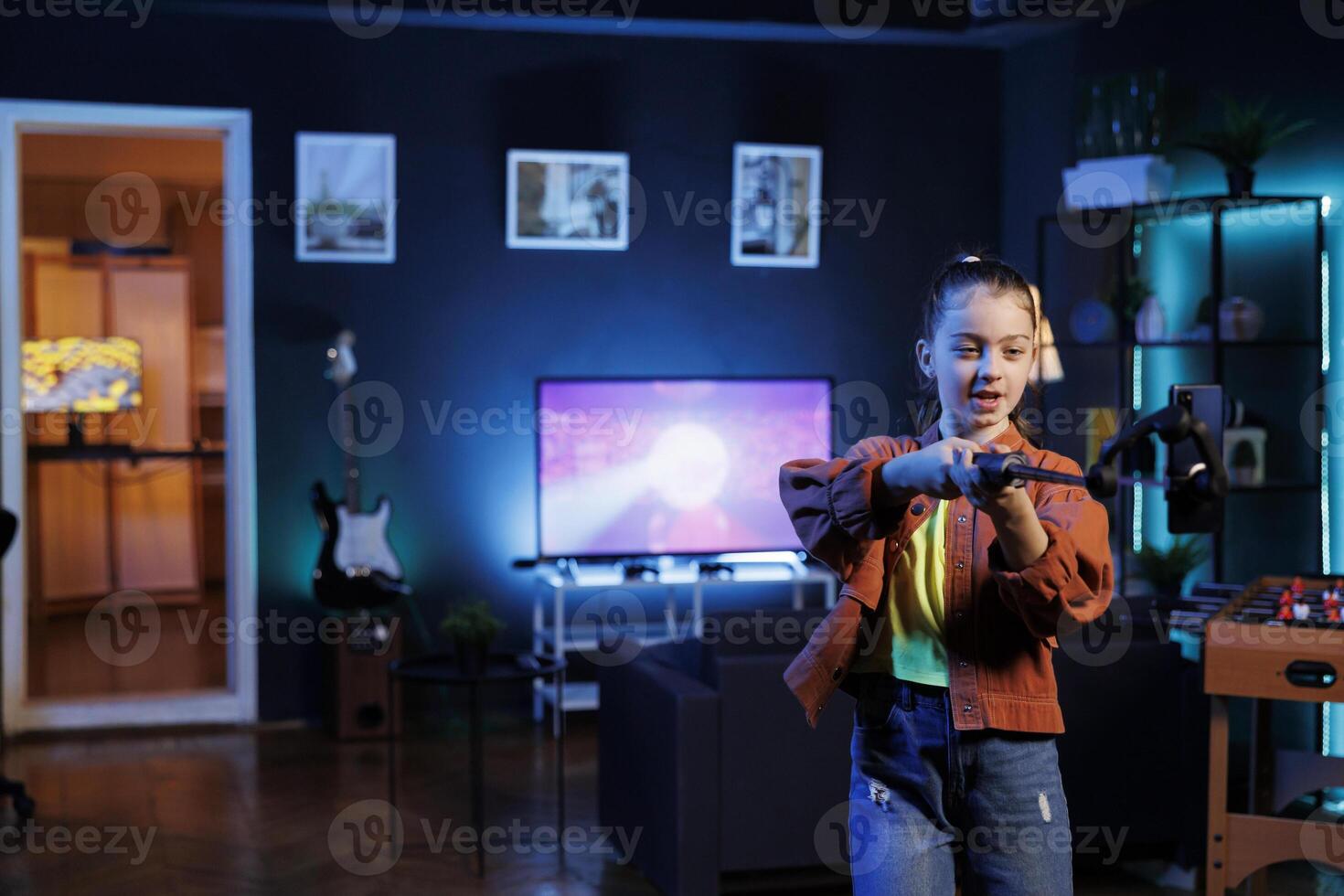 Kid taking part in viral dance trend after seeing favorite celebrities doing it, filming with cellphone on selfie stick. Girl does trendy challenge for online viewers, filming with phone photo