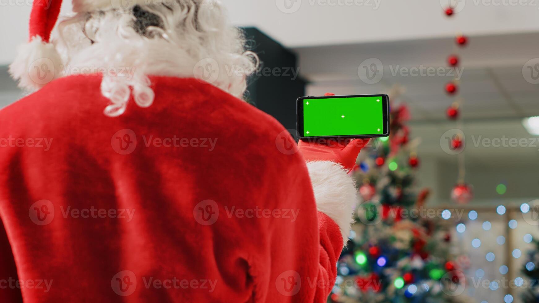 Employee in festive ornate shopping mall wearing Santa Claus suit holding green screen phone, taking a break from job shift and watching videos on chroma key screen, close up photo