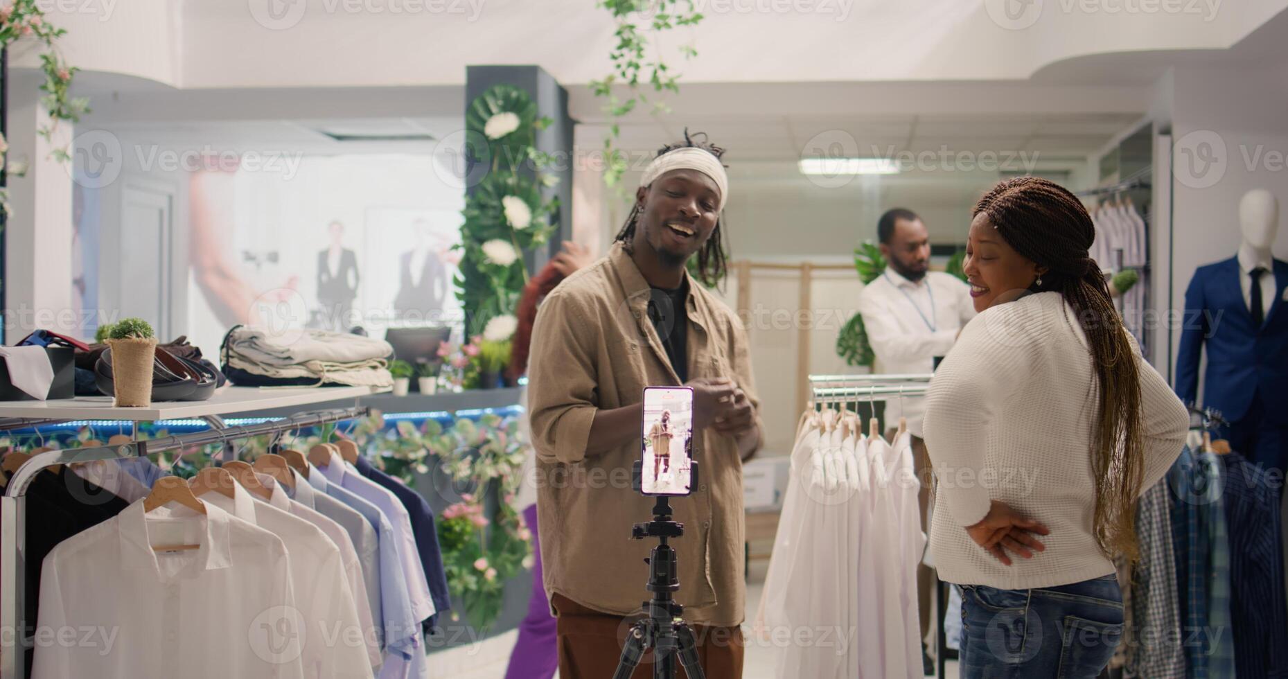 Internet star using smartphone to film couple daily vlog in clothing store with elegant assortment of clothes. Influencer and his girlfriend showing followers new collection of attire garments photo
