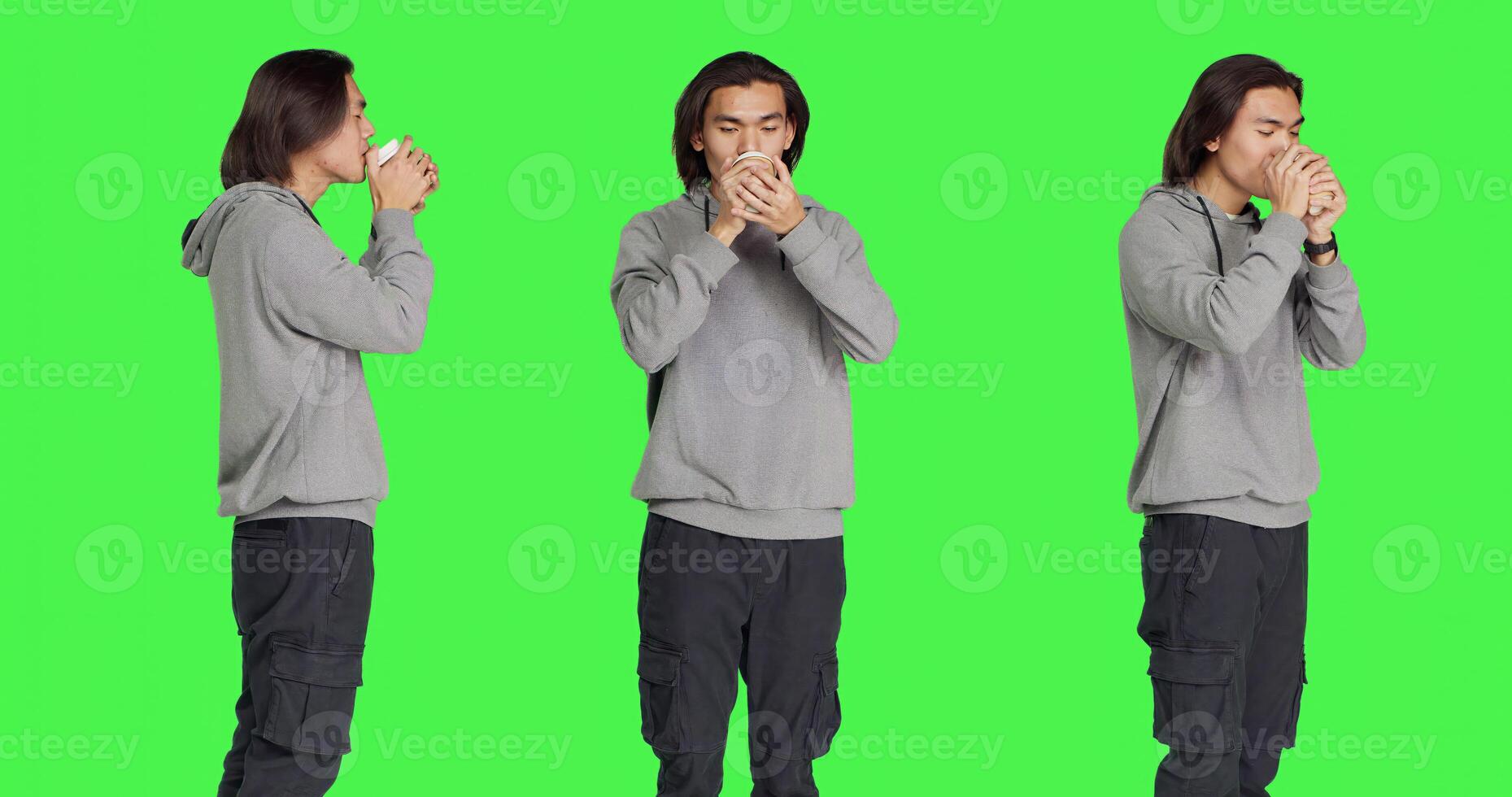 Person drinking cup of coffee on camera while standing in front of greenscreen template, enjoying caffeine refreshment. Young asian man holding hot beverage and feeling pleased. photo